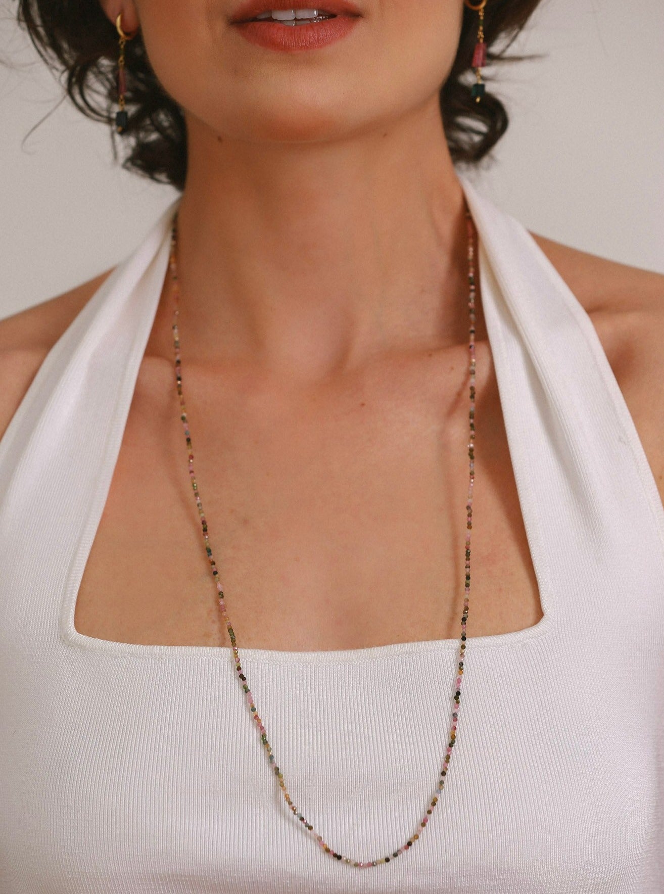 Modelo branca usando um colar de turmalina melancia natural todo colorido medindo 2mm cada pedrinha com um cumprimento de 80cm sendo usado com uma volta longo ao redor do pescoço com uma blusa branca e um brinco longo de turmalinas naturais.