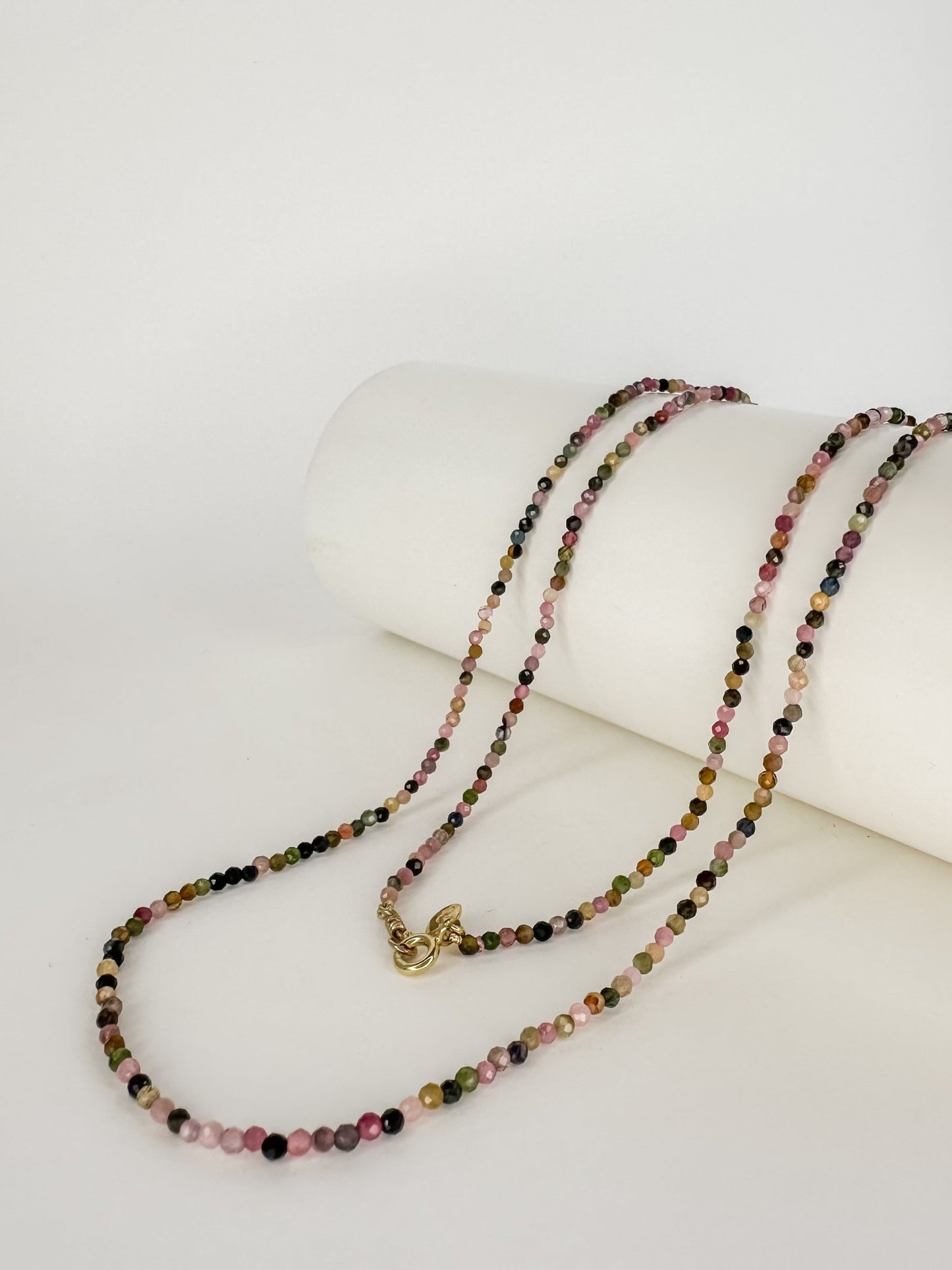 Colar Rasa de Turmalina melancia todo colorido aleatoriamente com pedrinhas de 2mm apoiado em um cilindo branco e fecho boia em prata com banho de ouro e fundo branco.