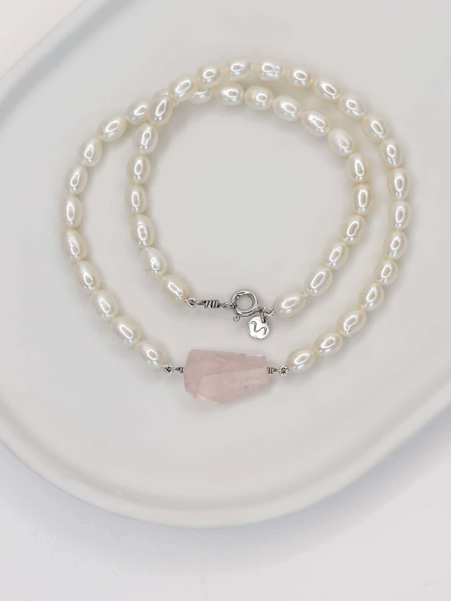 Colar de pérolas de modelo choker com fecho boia em prata com banho de ouro branco e detalhe no centro de um quartzo rosa de tamanho médio fixado por fio de prata com lapidação geométrica artesanal, apoiado em cerâmica branca com fundo branco.