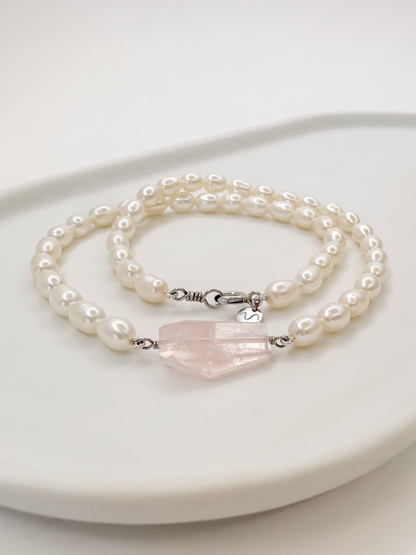 Colar de pérolas de modelo choker com fecho boia em prata com banho de ouro branco e detalhe no centro de um quartzo rosa de tamanho médio fixado por fio de prata com lapidação geométrica artesanal, apoiado em cerâmica branca com fundo branco.