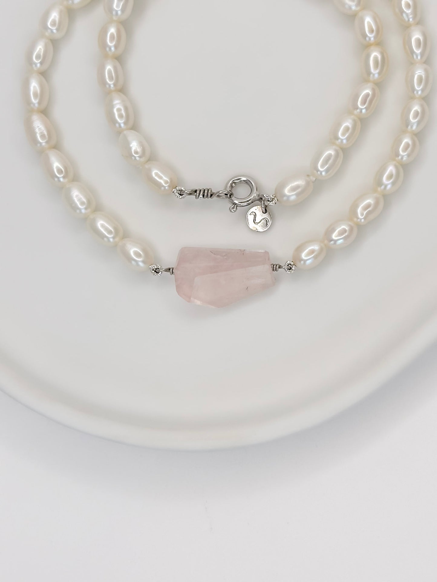 Colar de pérolas de modelo choker com fecho boia em prata com banho de ouro branco e detalhe no centro de um quartzo rosa de tamanho médio fixado por fio de prata com lapidação geométrica artesanal, apoiado em cerâmica branca com fundo branco.