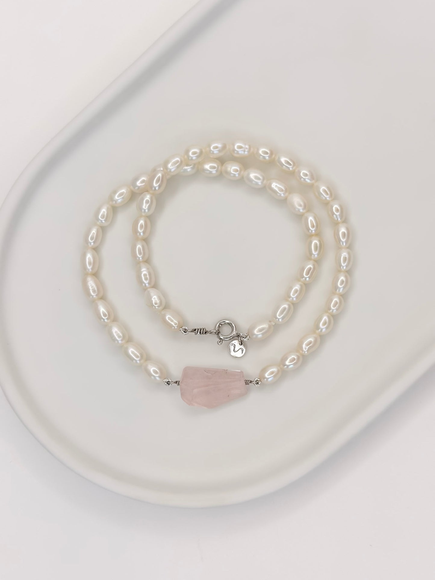 Colar de pérolas de modelo choker com fecho boia em prata com banho de ouro branco e detalhe no centro de um quartzo rosa de tamanho médio fixado por fio de prata com lapidação geométrica artesanal, apoiado em cerâmica branca com fundo branco.