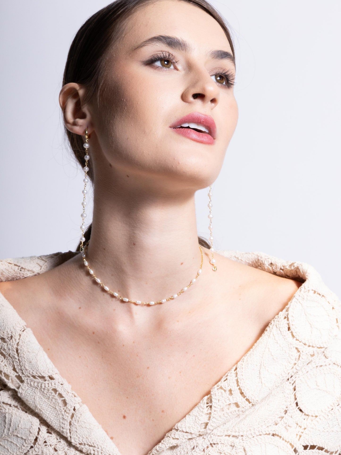 Modelo de pele branca usando colhar choker de 35 cm feito em prata com banho de ouro e com pérolas naturais de agua doce vestindo blazer de renda e brincos longos de perolas naturais com fundo branco.