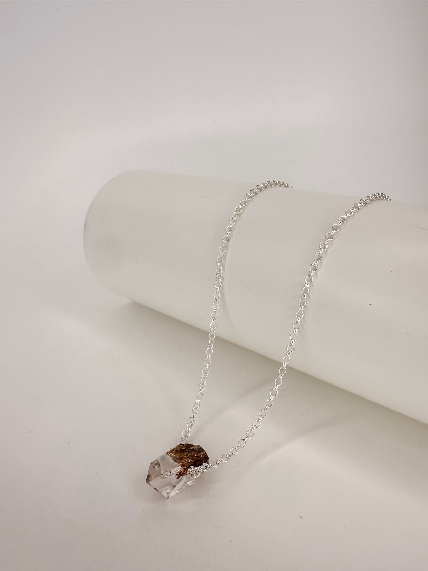 Colar em corrente portuguesa de prata apoiado em cilindro com uma ponta do cristal natural quartzo lodolita com fundo branco.