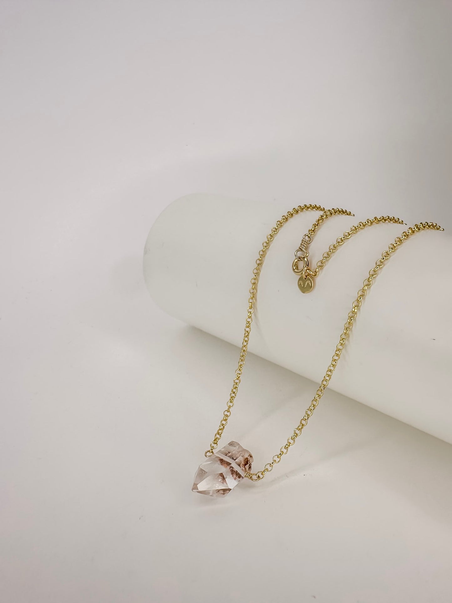 Colar em corrente portuguesa de prata com banho de ouro apoiado em cilindro com uma ponta do cristal natural quartzo lodolita com fundo branco.