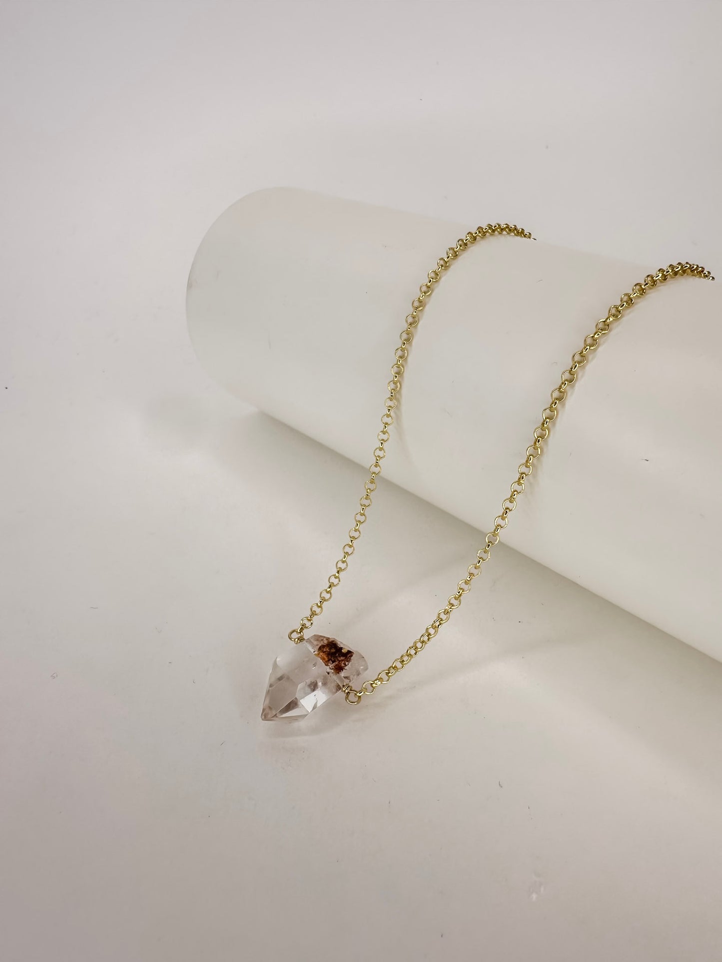 Colar em corrente portuguesa de prata com banho de ouro apoiado em cilindro com uma ponta do cristal natural quartzo lodolita com fundo branco.