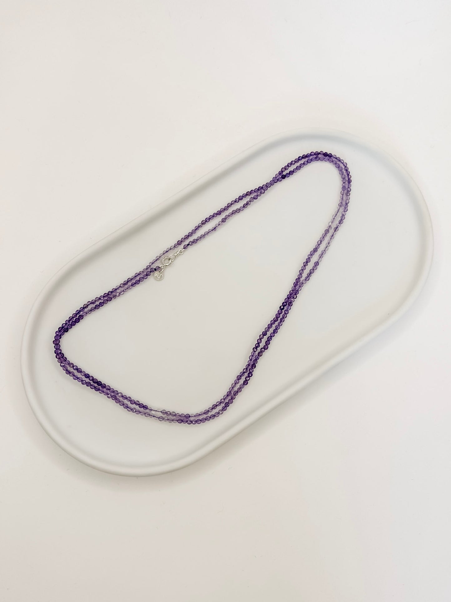 Colar feito com esferas de cristal natural ametista lapidados de aproximadamente 2mm com cumprimento de 80 cm apoiado em bandeja de ceramica branca e fundo branco.