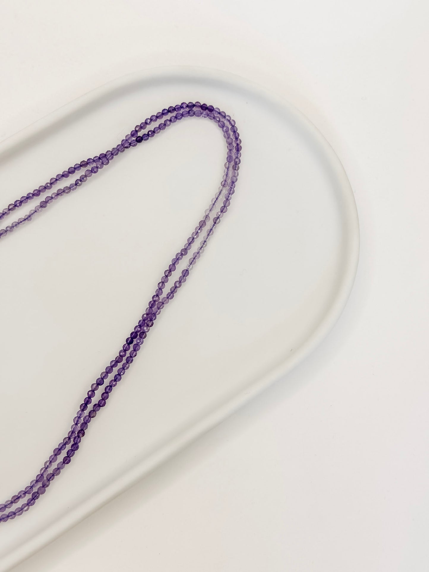 Colar feito com esferas de cristal natural ametista lapidados de aproximadamente 2mm com cumprimento de 80 cm apoiado em bandeja de ceramica branca e fundo branco.