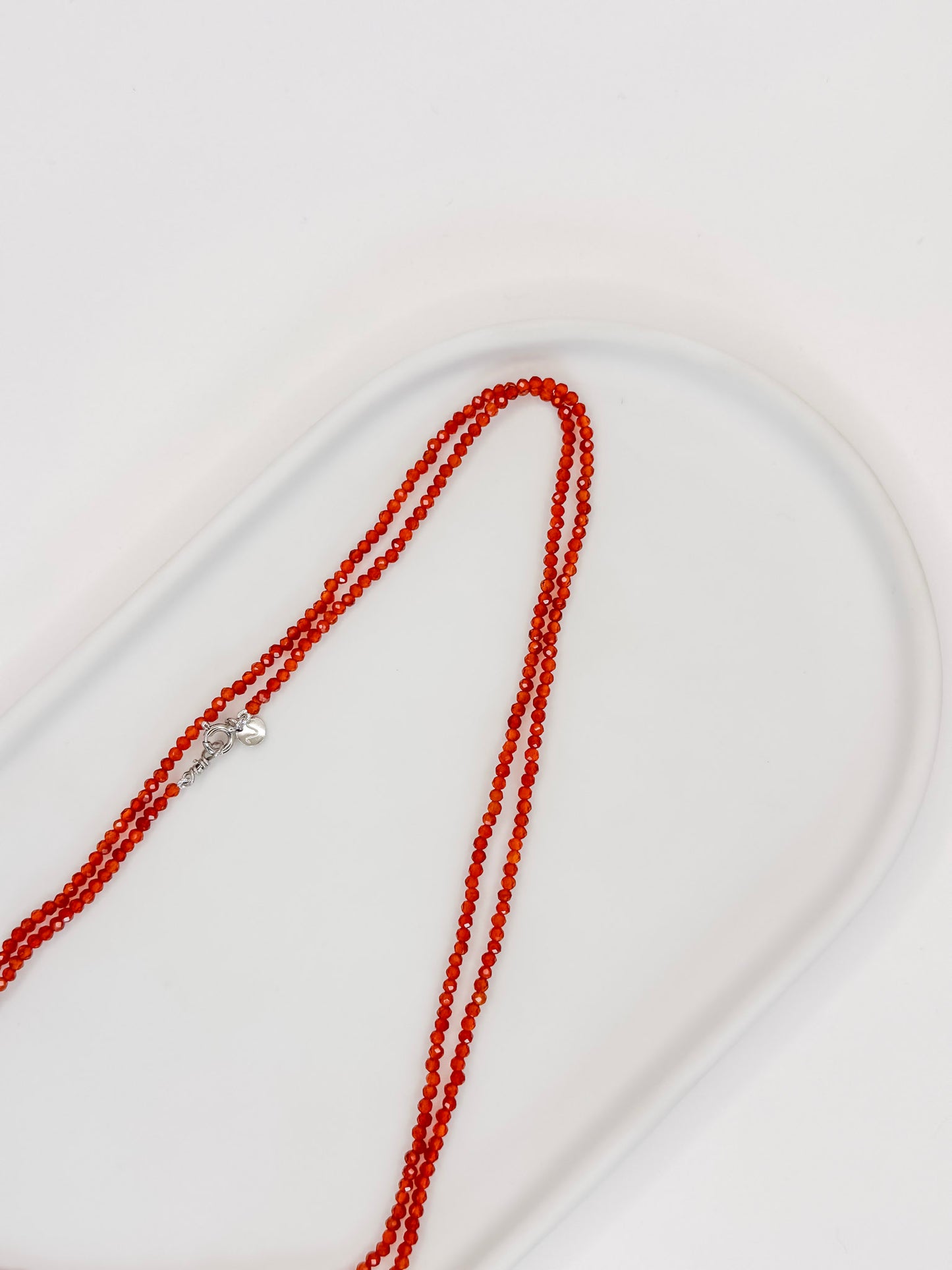 Foto Colar de cristais Cornalina Natural com aproximadamente 80 cm e fecho boia em prata com banho de ouro branco, apoiado em bandeja de cerâmica branca com fundo branco.