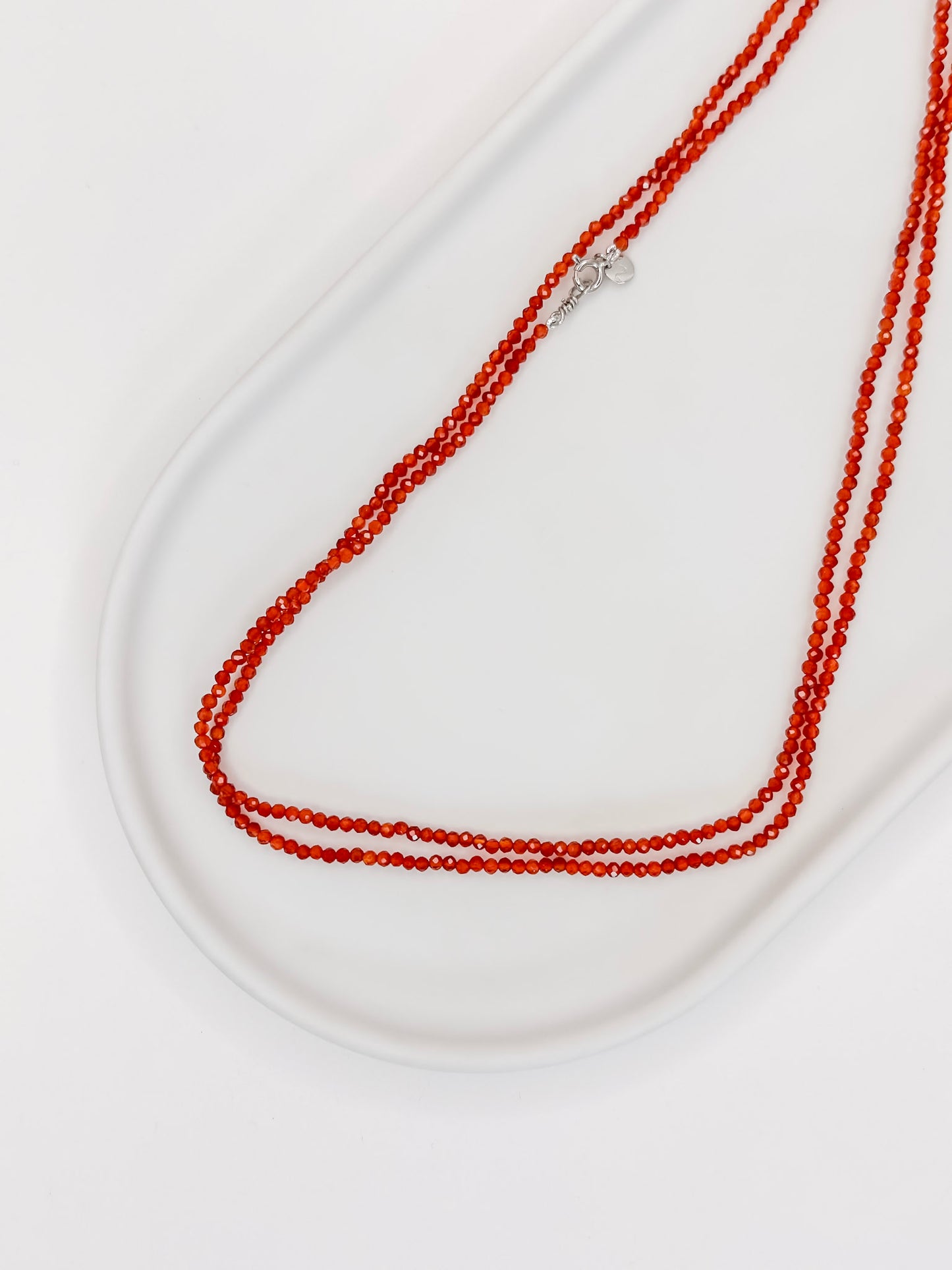 Foto Colar de cristais Cornalina Natural com aproximadamente 80 cm e fecho boia em prata com banho de ouro branco, apoiado em bandeja de cerâmica branca com fundo branco.
