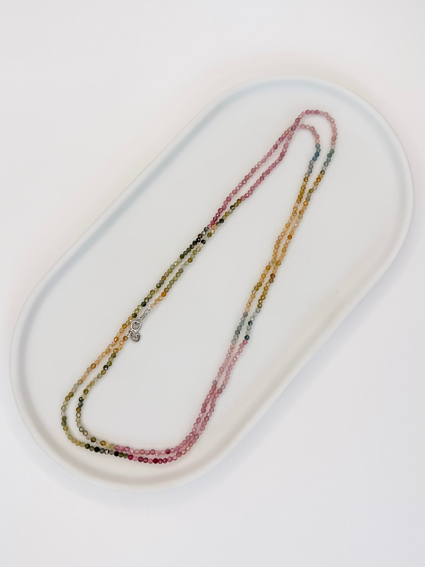 Colar produzido com esferas lapidadas de turmalinas melancia naturais medindo aproximadamente 2mm cada, com um cumprimento de 80 cm com fecho boia em prata, apoiado em uma bandeja branca com fundo branco.