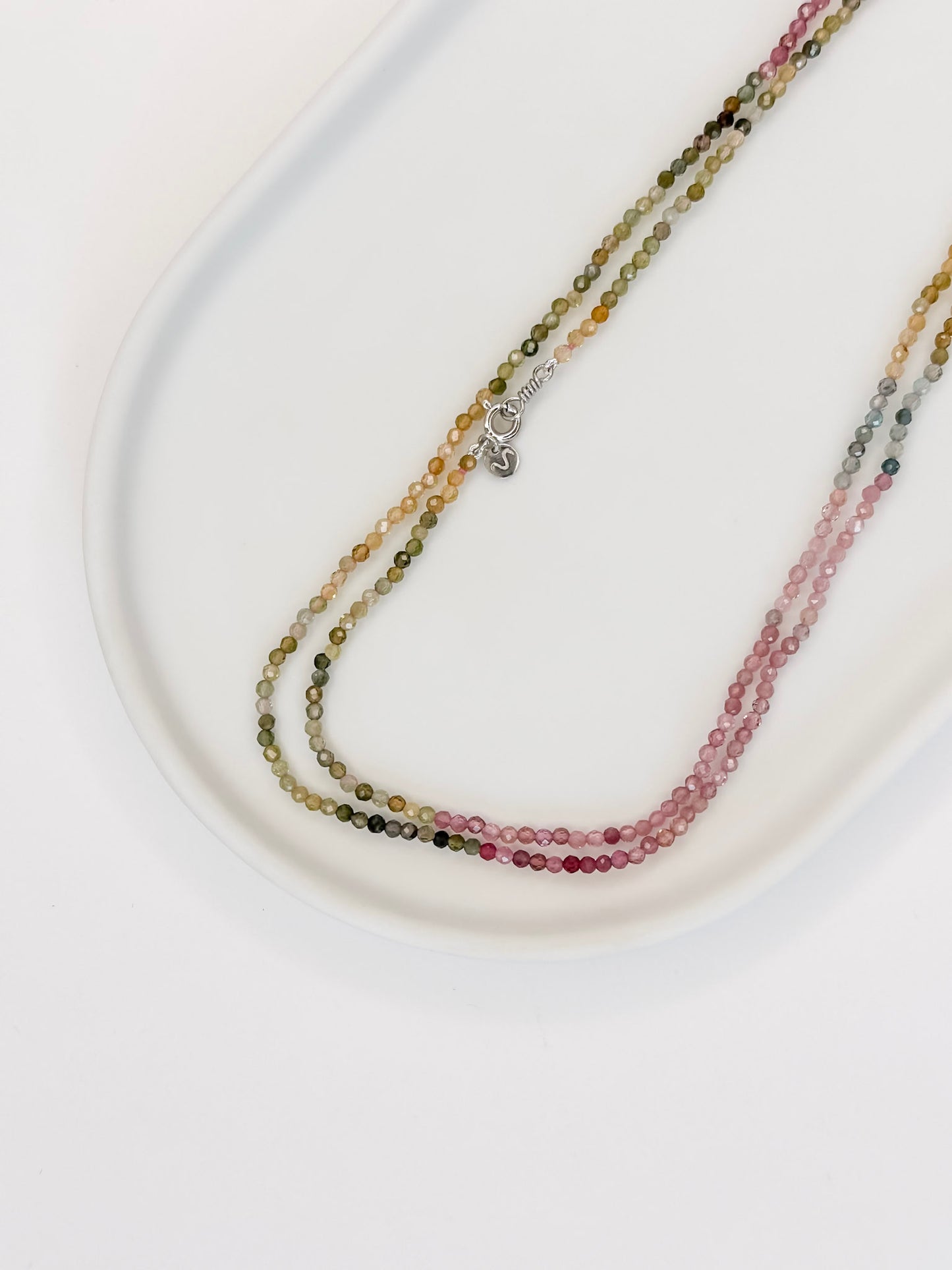 Colar produzido com esferas lapidadas de turmalinas melancia naturais medindo aproximadamente 2mm cada, com um cumprimento de 80 cm com fecho boia em prata, apoiado em uma bandeja branca com fundo branco.