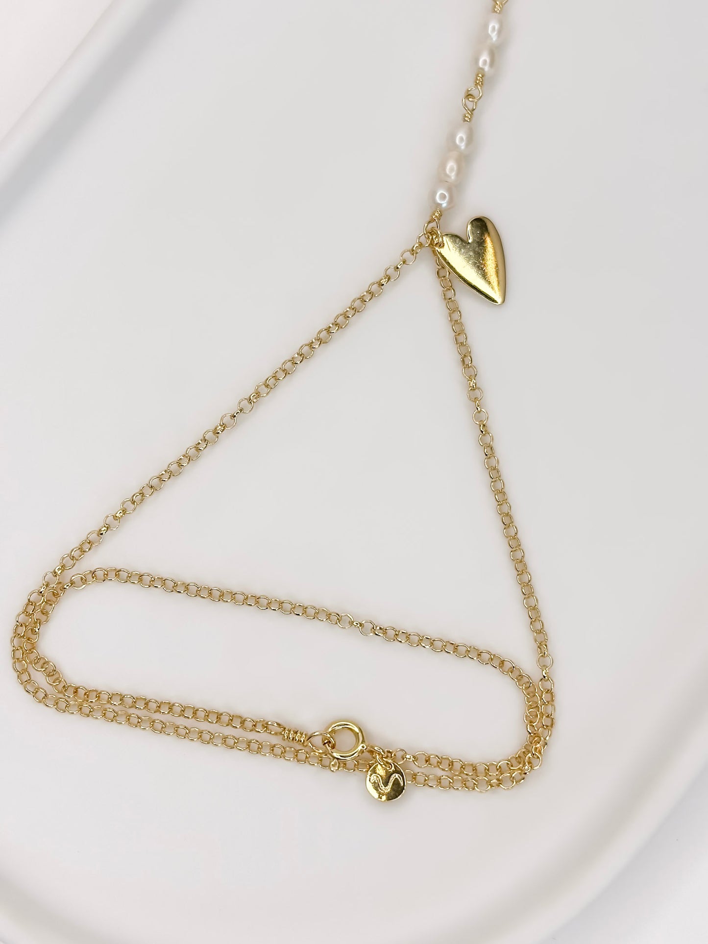 Detalhe da pulseira de mão feita com corrente portuguesa em prata 925 com banho de ouro 18k, pérolas naturais e um pingente em formato de coração apoiada em cerâmica branca com fundo branco.