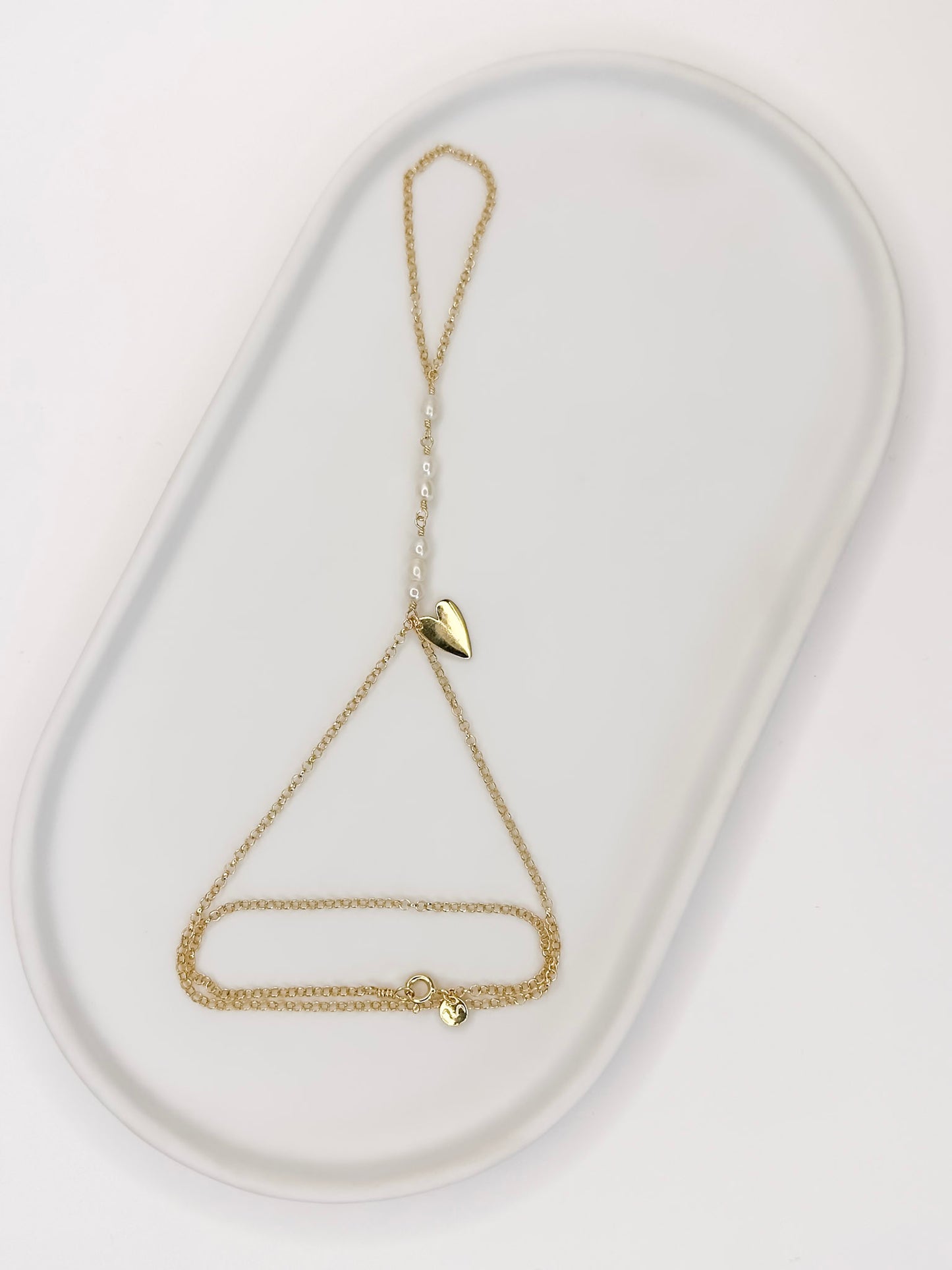 Pulseira de mão feita com corrente portuguesa em prata 925 com banho de ouro 18k, pérolas naturais e um pingente em formato de coração apoiada em cerâmica branca com fundo branco.