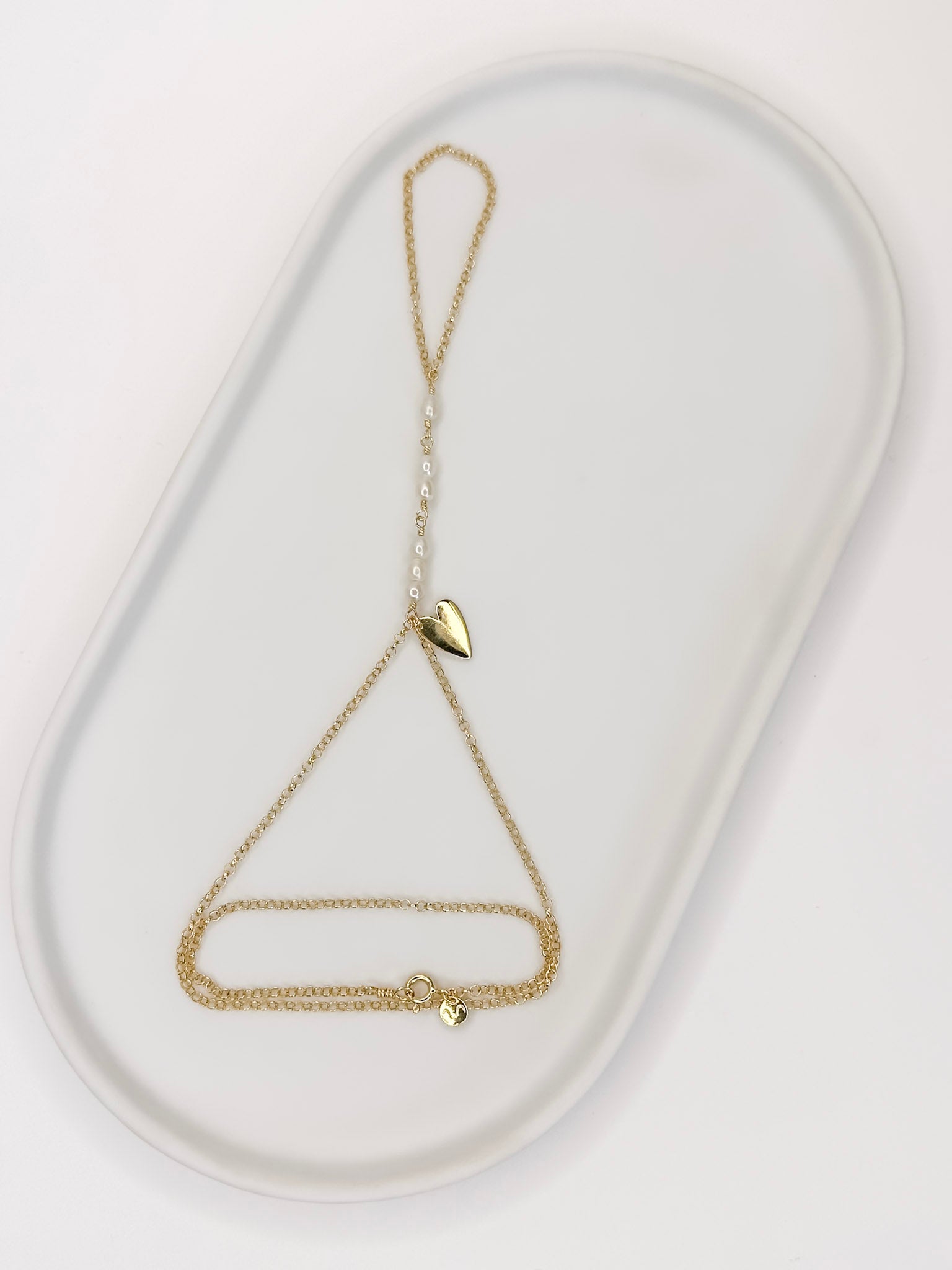 Pulseira de mão feita com corrente portuguesa em prata 925 com banho de ouro 18k, pérolas naturais e um pingente em formato de coração apoiada em cerâmica branca com fundo branco.