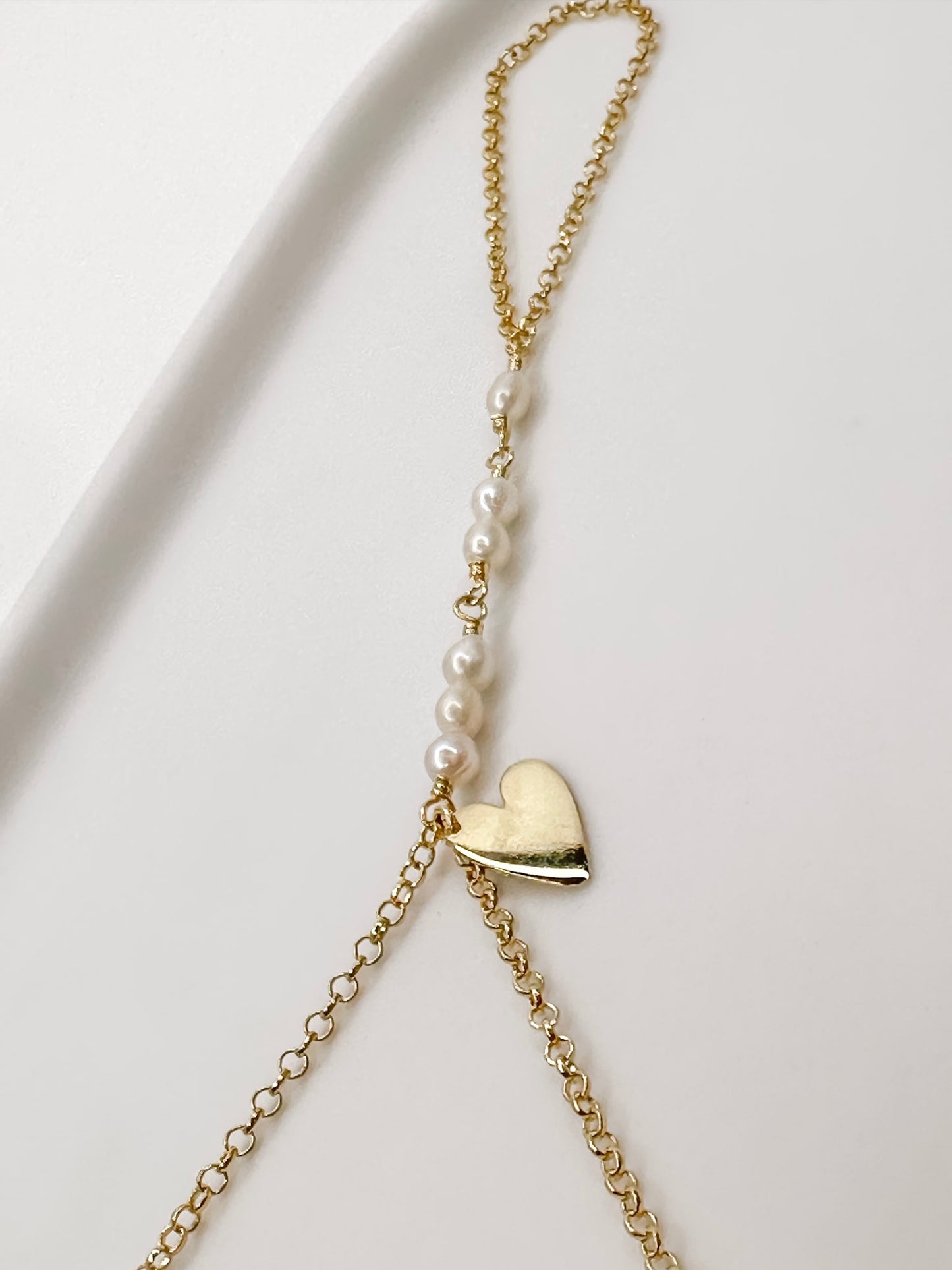 Detalhe da pulseira de mão feita com corrente portuguesa em prata 925 com banho de ouro 18k, pérolas naturais e um pingente em formato de coração apoiada em cerâmica branca com fundo branco.