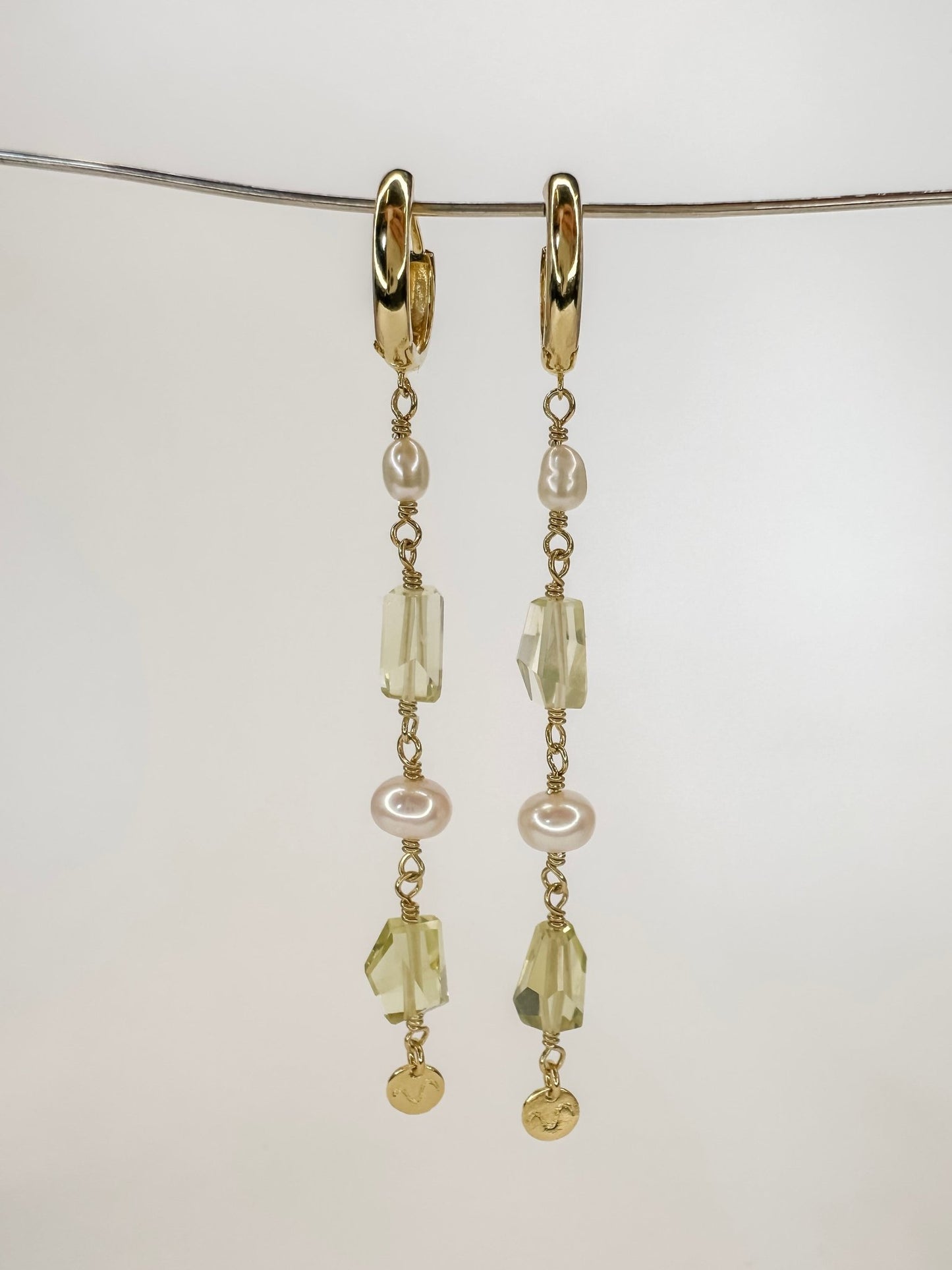 Brinco longo de argola click em prata com banho de ouro com duas pedras de quartzo limao ou quartzo verde lapidadas e duas perolas tendo o cumprimento longo e com uma plaquinha pendurada no final, com fundo branco.