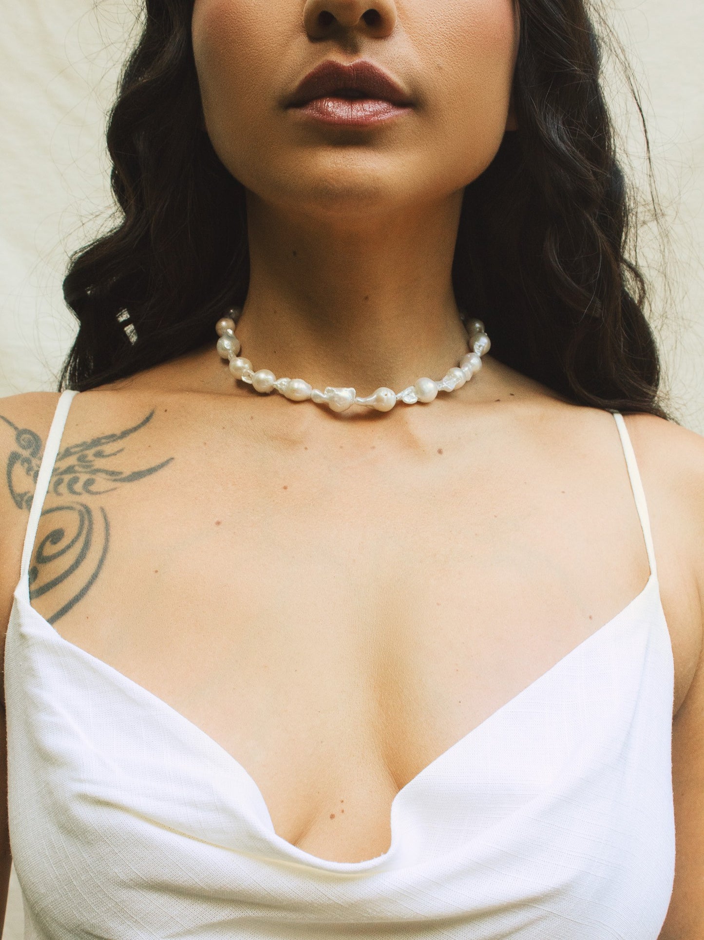 Colar estilo gargantilha com pérolas barrocas grandes vestidos em mulher morena om vestido branco e fundo claro.