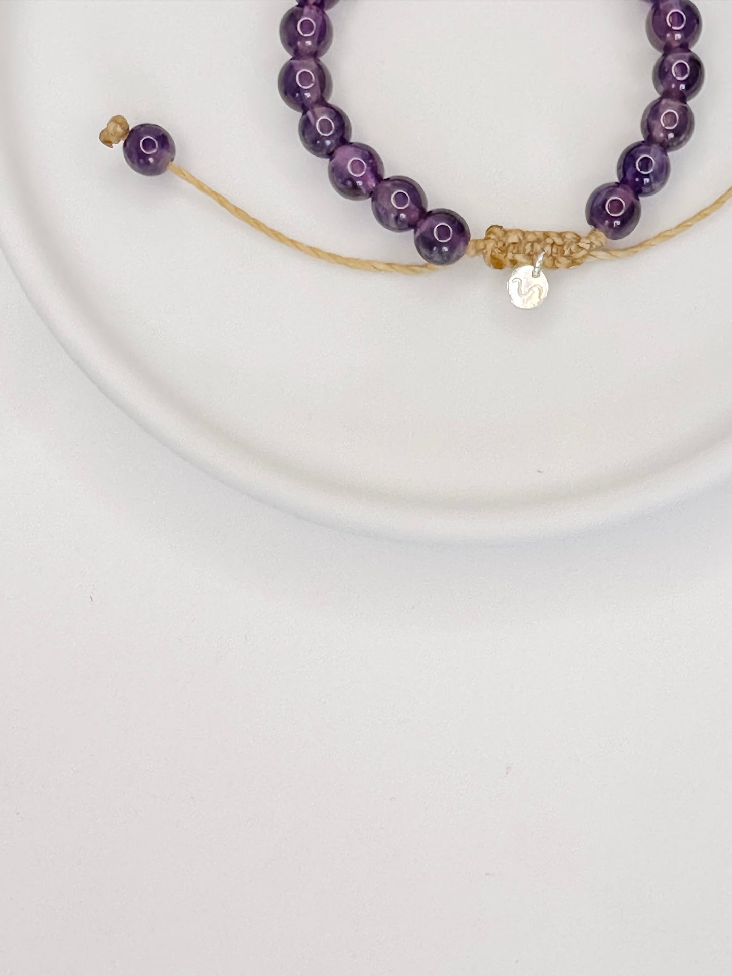 Pulseira feita com corda e esferas lisas de cristal ametista, tamanho unico, apoiada em ceramica branca com fundo branco.