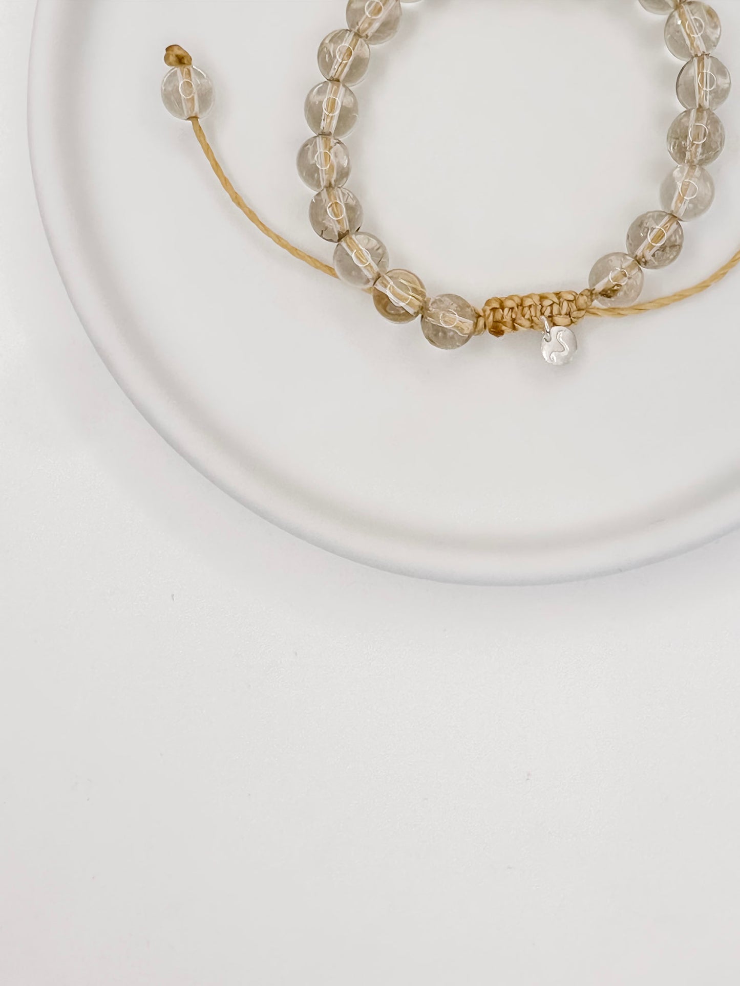 Pulseira de corda tamanho único com esferas de 6mm de quartzo rutilado natural apoiado em prato de cerâmica branco e fundo branco.