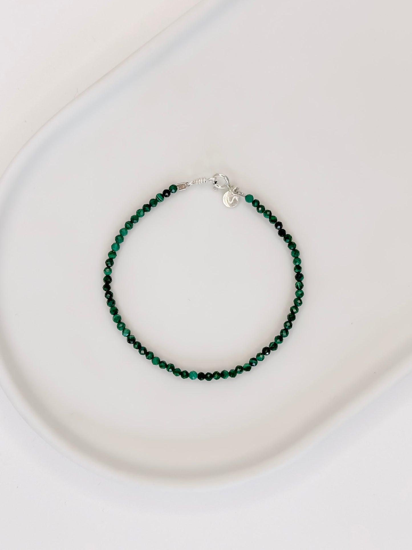 Pulseira com pedra malaquita natural com esferas lapidadas de aproximadamente 2mm apoiada em bandeja de ceramica branca com fundo branco.