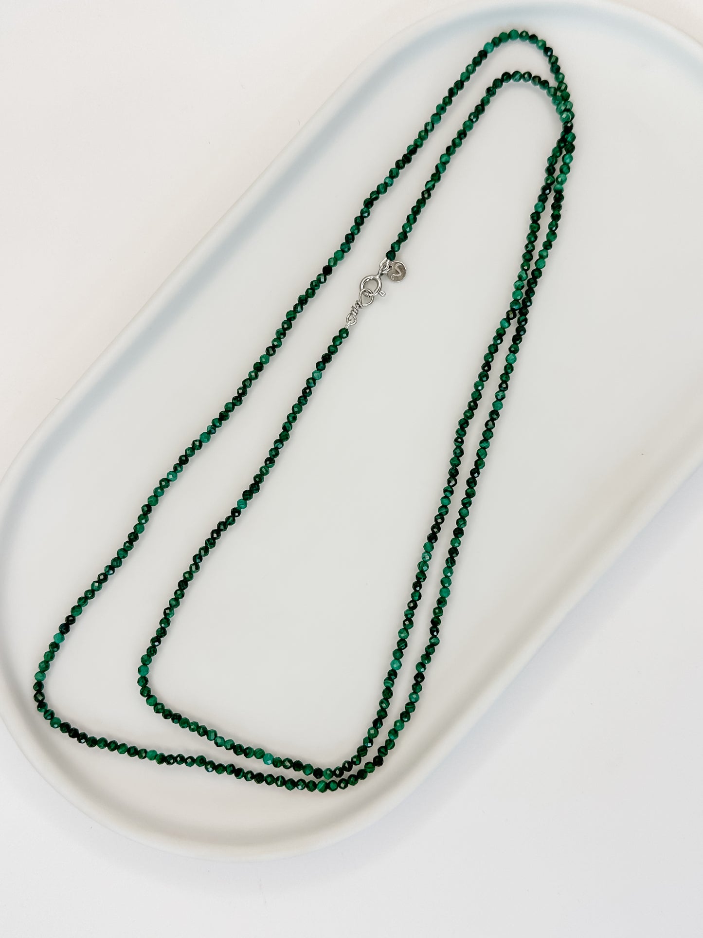 Colar de 80 cm produzido com esferas lapidadas de cristal malaquita medindo aproximadamente 2mm cada com fecho bóia em prata apoiado em cerâmica branca e fundo branco.