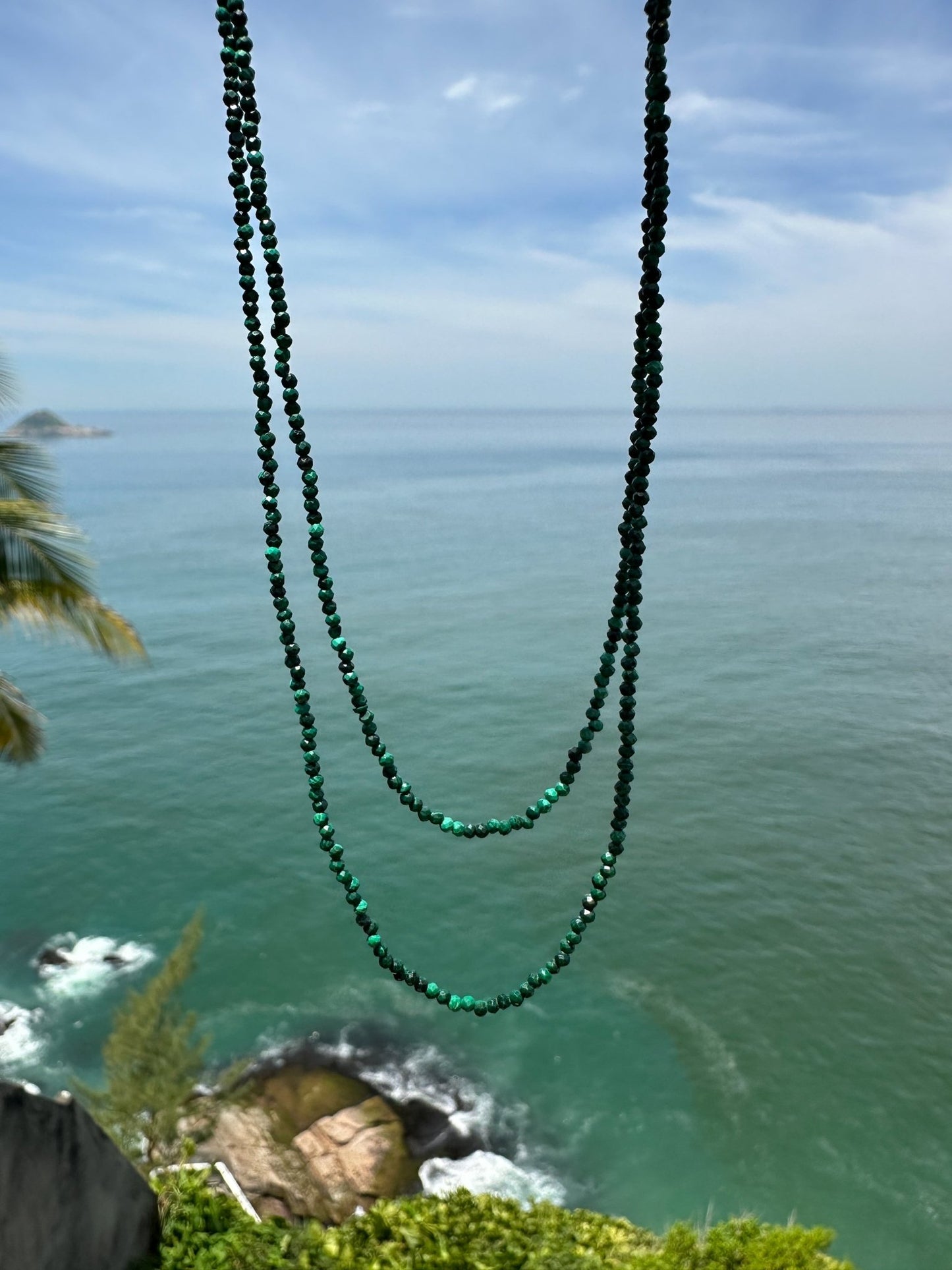 Colar de 80 cm produzido com esferas lapidadas de cristal malaquita medindo aproximadamente 2mm cada com fecho bóia em prata com fundo de horizonte do mar com rochas e vegetação.