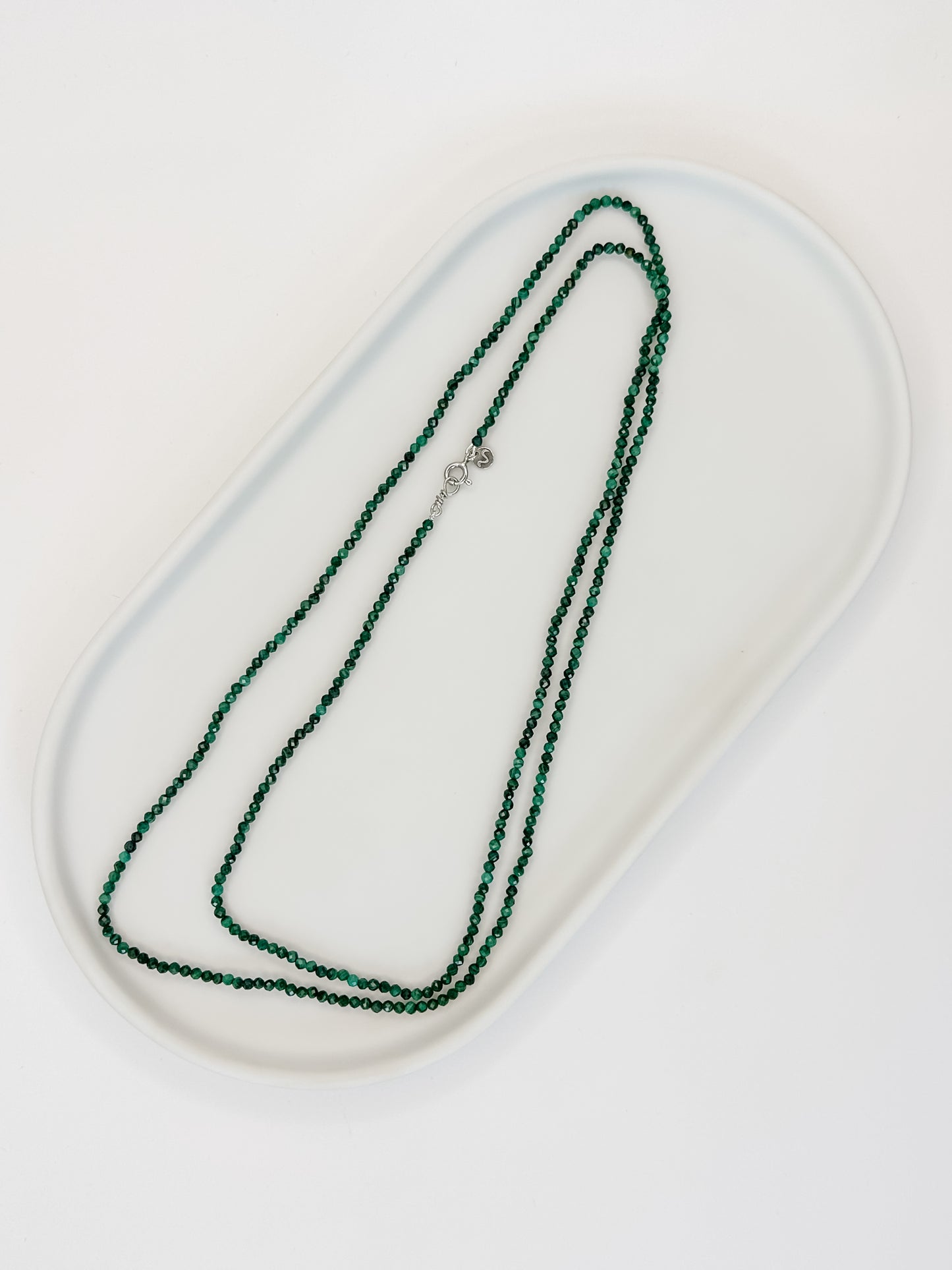 Colar de 80 cm produzido com esferas lapidadas de cristal malaquita medindo aproximadamente 2mm cada com fecho bóia em prata apoiado em cerâmica branca e fundo branco.