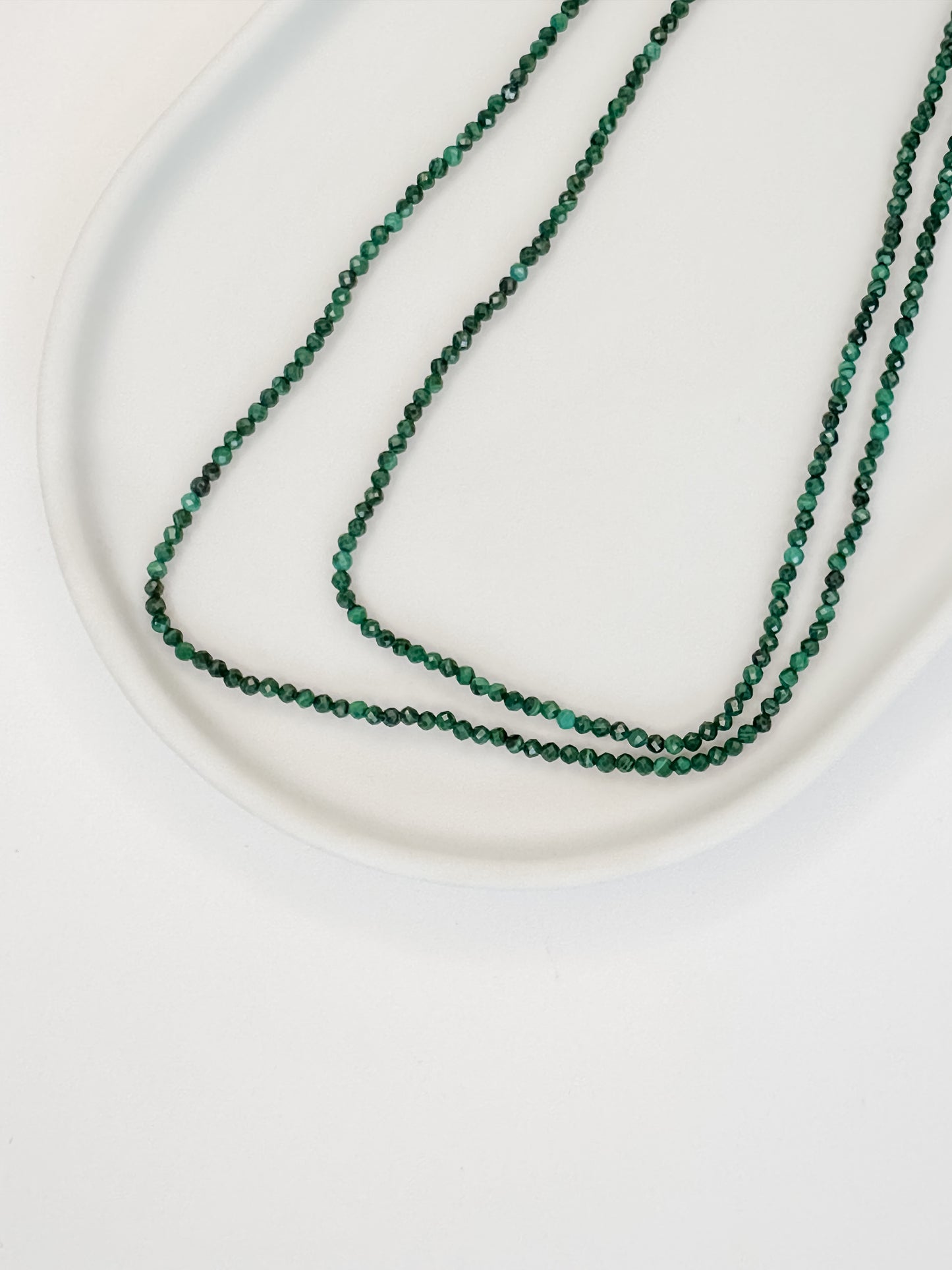 Colar de 80 cm produzido com esferas lapidadas de cristal malaquita medindo aproximadamente 2mm cada com fecho bóia em prata apoiado em cerâmica branca e fundo branco.