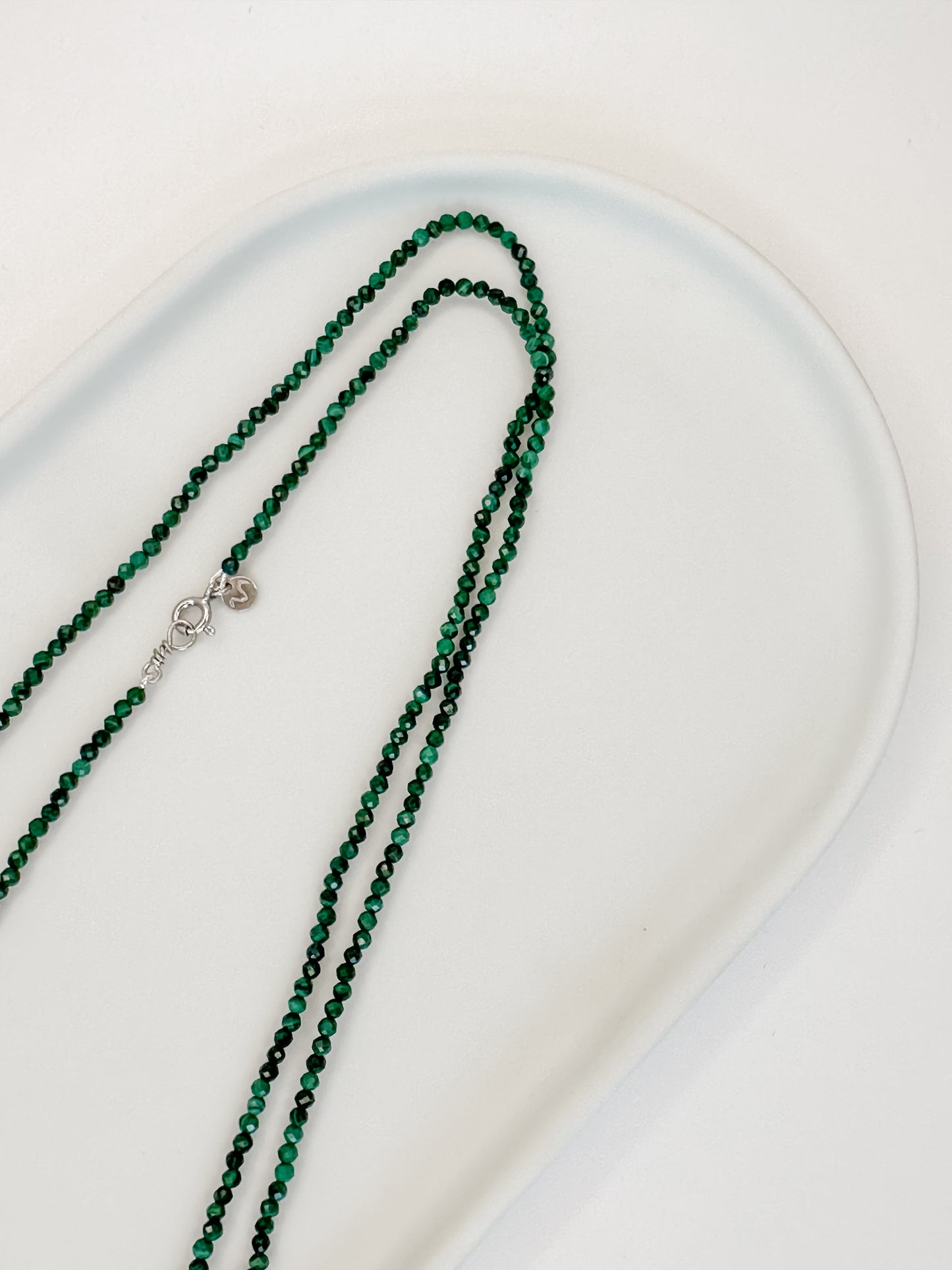 Colar de 80 cm produzido com esferas lapidadas de cristal malaquita medindo aproximadamente 2mm cada com fecho bóia em prata apoiado em cerâmica branca e fundo branco.