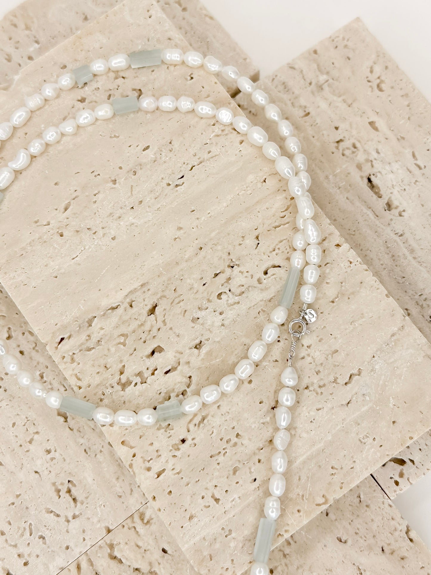 Detalhe Colar longo com perolas ovais intercalado com pedras naturais água marinha brasileiras, apoiado em marmore com fundo branco.
