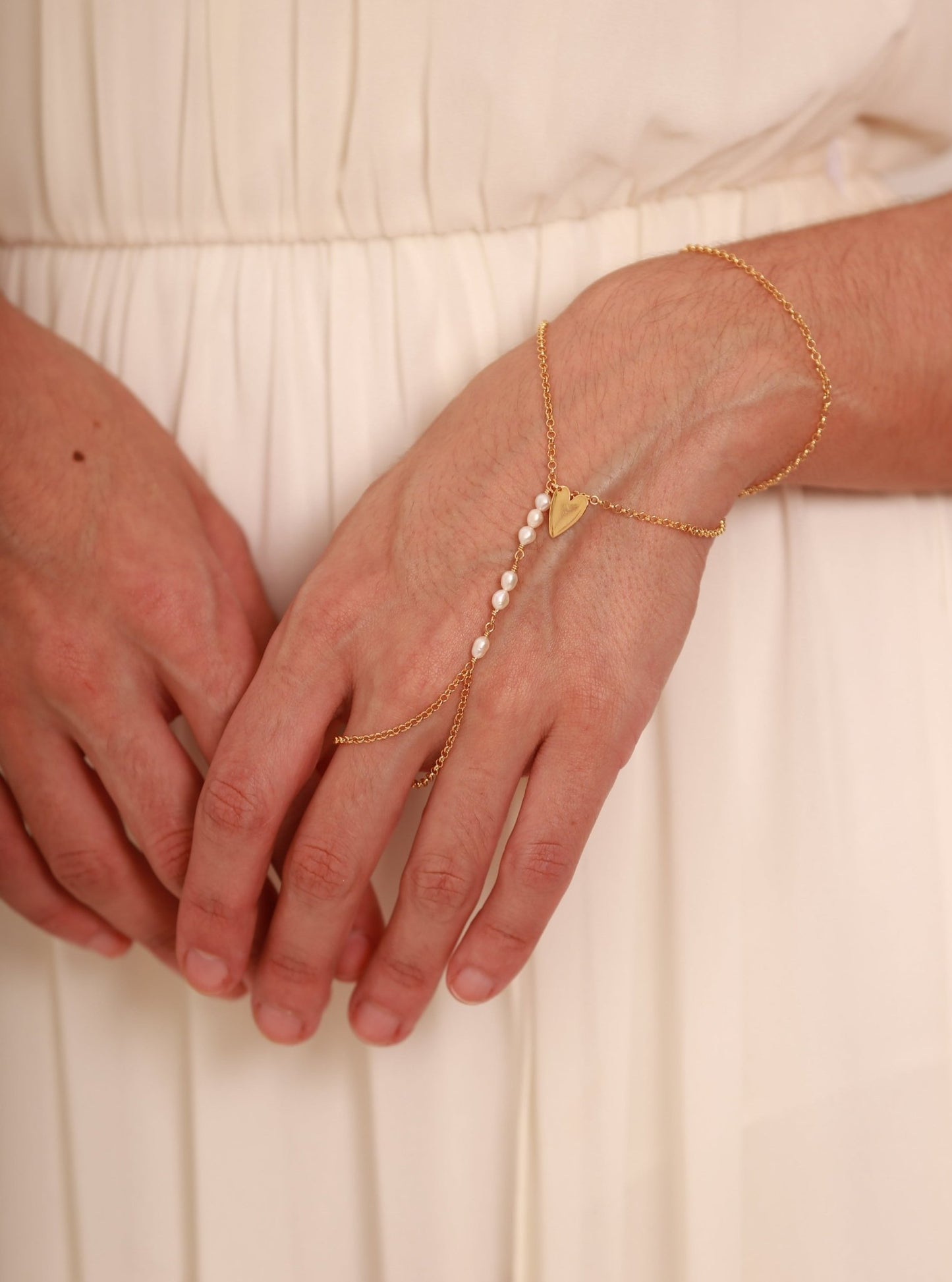 Detalhe Modelo vestindo Pulseira de mão com corrente portuguesa em prata 925 com banho de ouro 18k com mini pérolas e pingente coração e fundo branco.