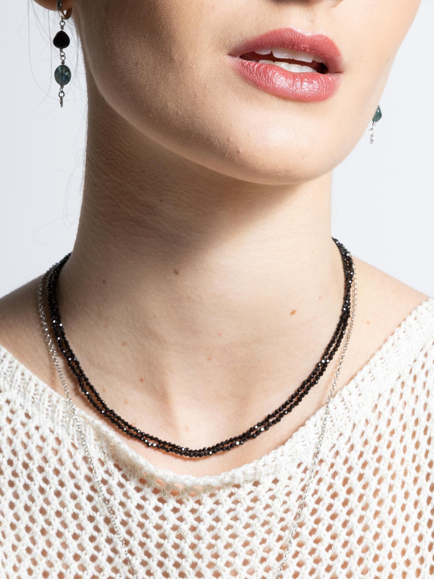 Modelo vestindo Colar de duas voltas produzido com esferas facetadas de espinelio natural medindo aproximadamente 2mm cada, com fecho em prata 925, blusa em tricot bege e brincos com pedras lisas azul e preto e fundo branco.