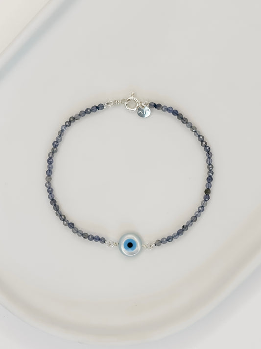 Pulseira feita com esferas lapidadas de 2mm do cristal Iolita com tom azul fechado, com um olho em madrepérola fixado no centro e fecho bóia em prata apoiada em bandeja branca com fundo branco. 