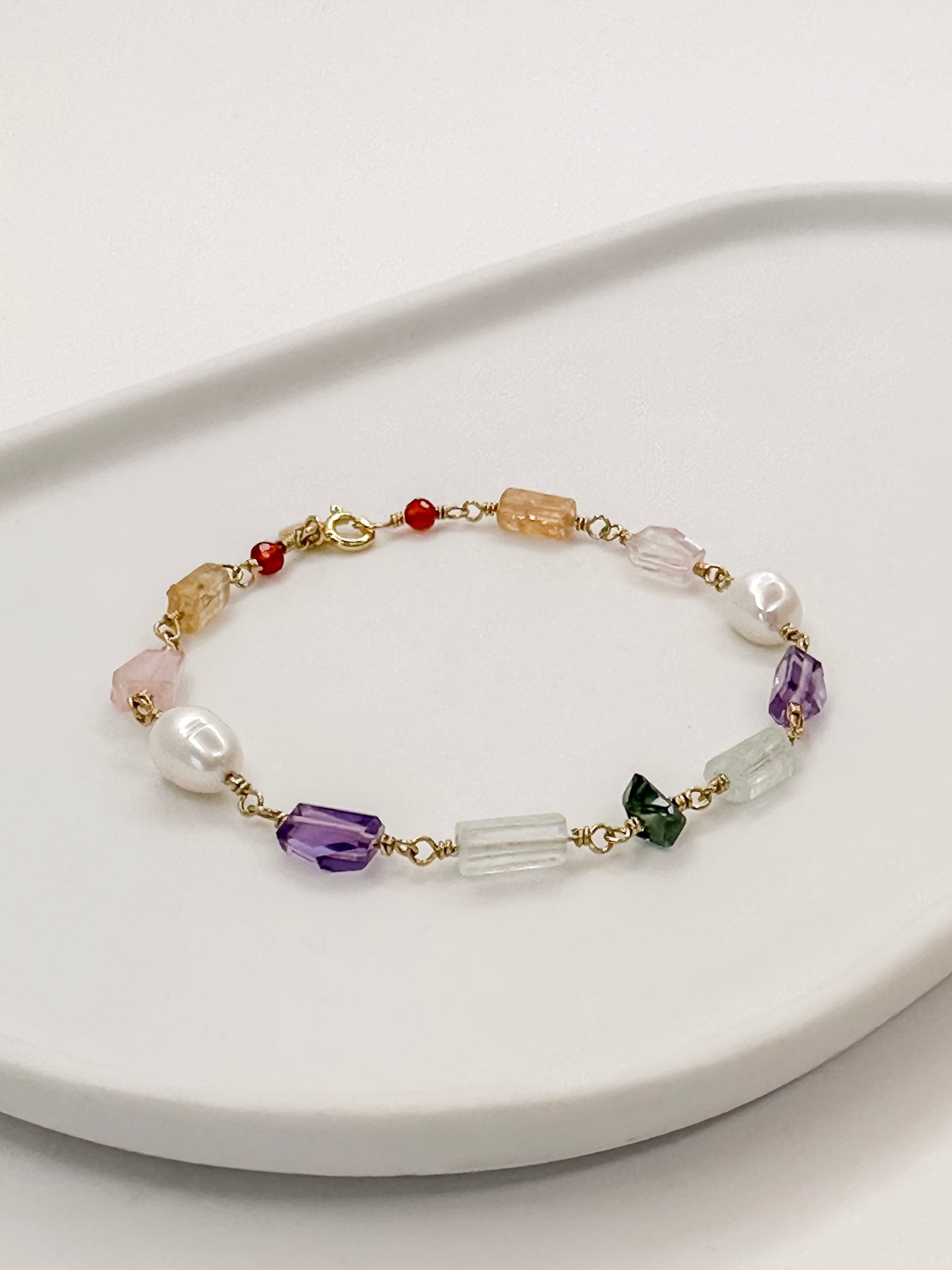 Pulseira feita a mao com perola e cristais naturais com cornalina, topazio imperial, quartzo rosa, ametista, agua marinha e turmalina apoiada em bandeja branca com fundo branco.