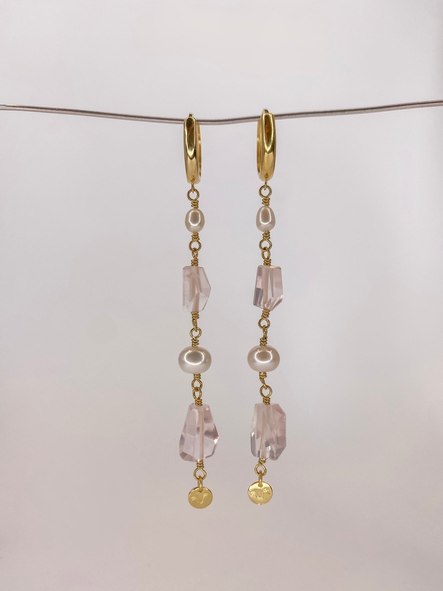 Brinco de argola click em prata com banho de ouro com duas perolas naturais e duas pedras de cristais quartzo rosa naturais lapidadas manualmente sendo um brinco longo com detalhe de placa da marca na ponta, pendurado em um fio de metal com fundo branco.