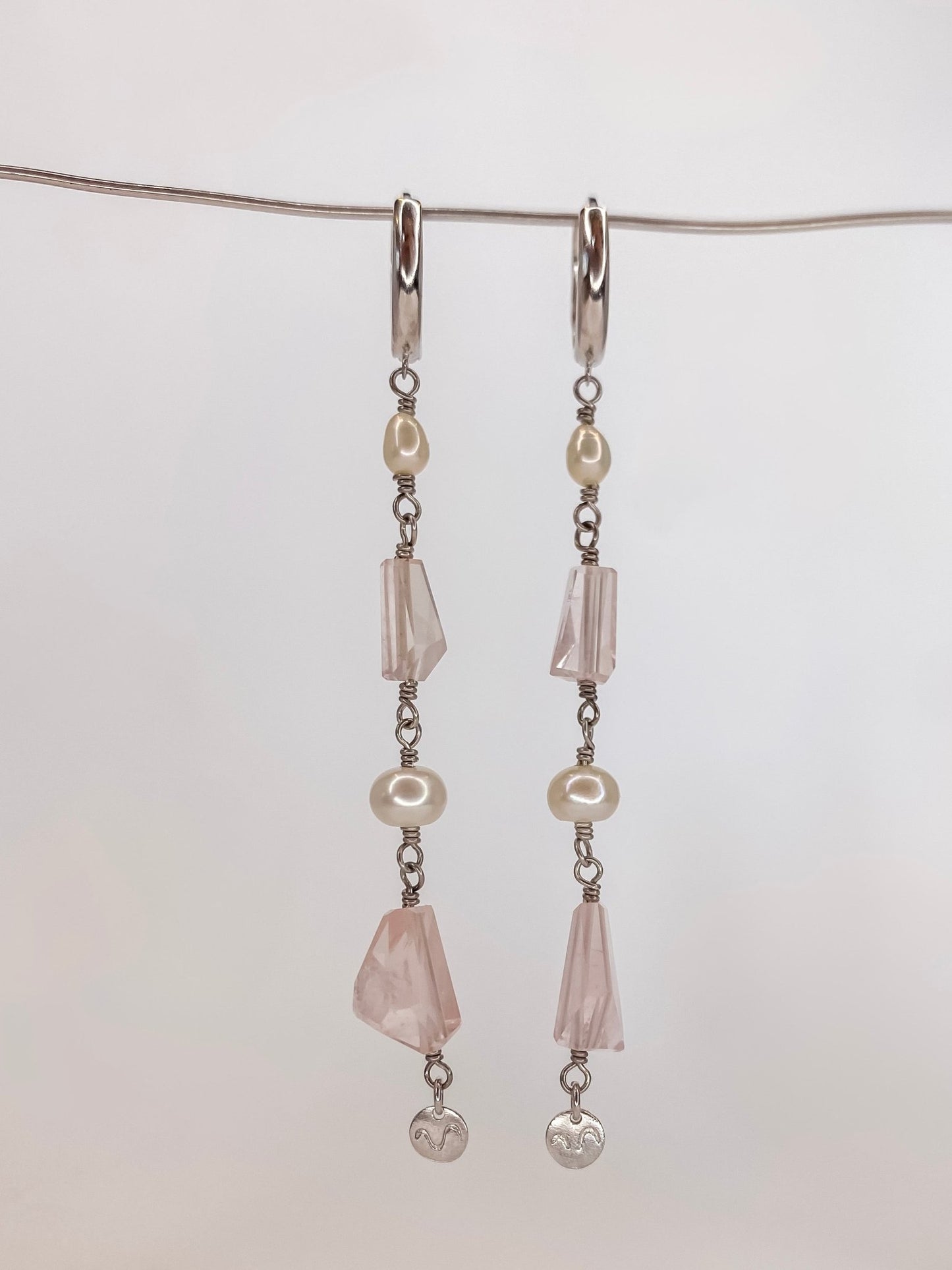 Brinco de argola click em prata com banho de ouro branco com duas perolas naturais e duas pedras de cristais quartzo rosa naturais lapidadas manualmente sendo um brinco longo com detalhe de placa da marca na ponta, pendurado em um fio de metal com fundo branco.