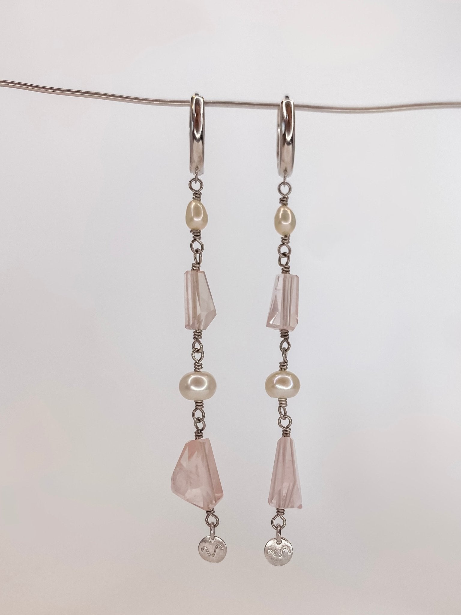 Brinco de argola click em prata com banho de ouro branco com duas perolas naturais e duas pedras de cristais quartzo rosa naturais lapidadas manualmente sendo um brinco longo com detalhe de placa da marca na ponta, pendurado em um fio de metal com fundo branco.