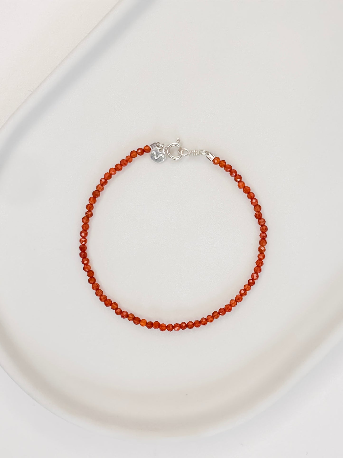 Pulseira em cristal natural vermelho Cornalina com esferas lapidadas de aproximadamente 2mm com fecho boia em prata, apoiada em bandeja de cerâmica branca e fundo branco.