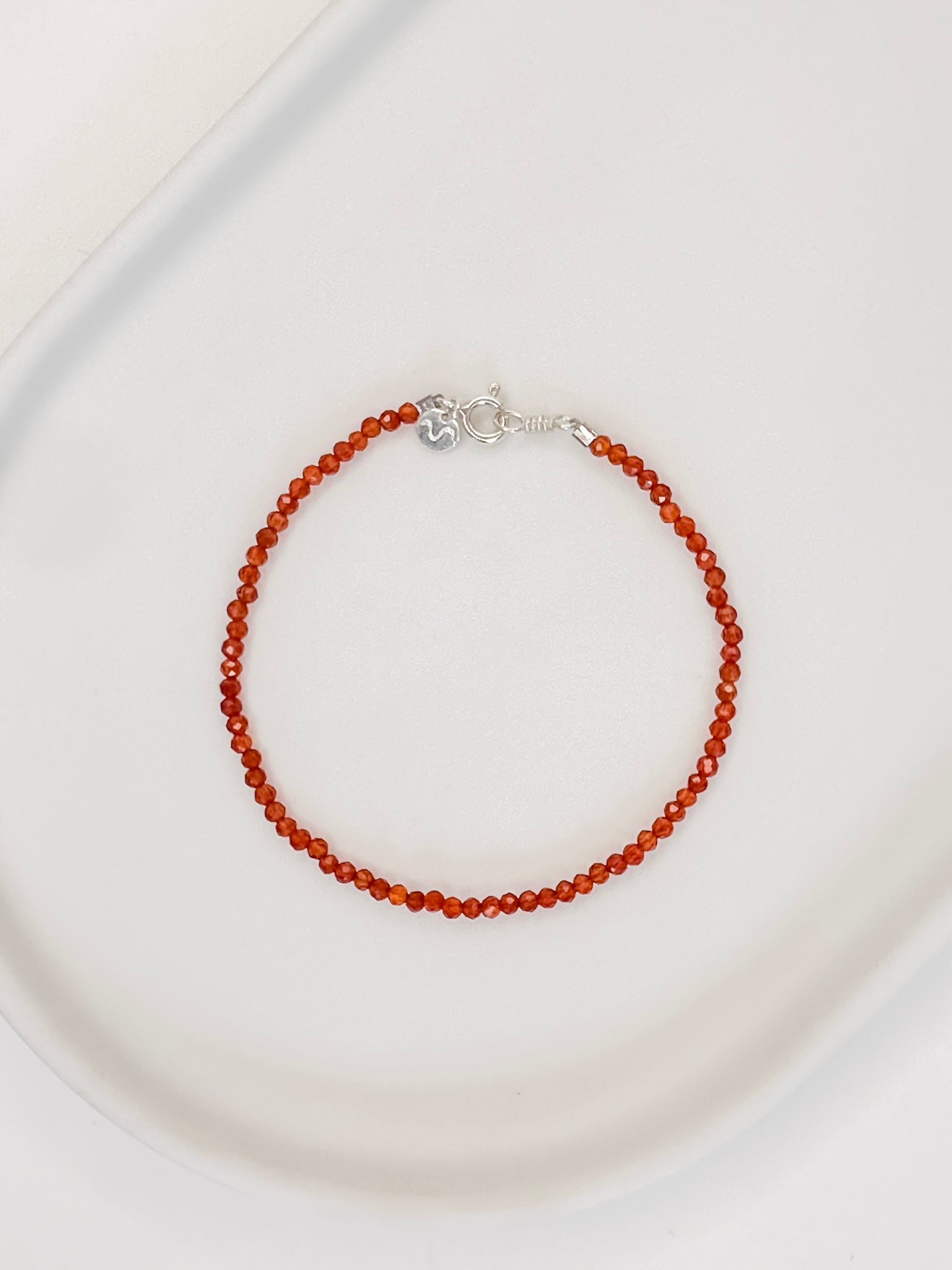 Pulseira em cristal natural vermelho Cornalina com esferas lapidadas de aproximadamente 2mm com fecho boia em prata, apoiada em bandeja de cerâmica branca e fundo branco.
