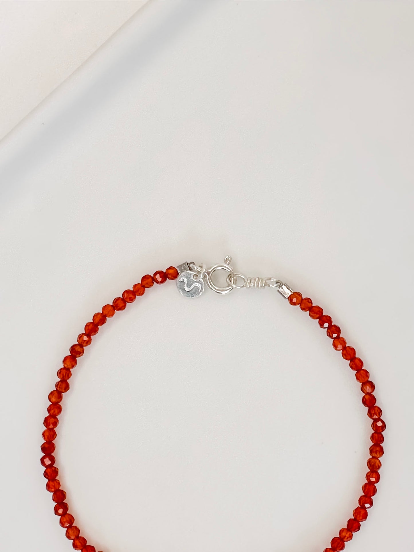 Pulseira em cristal natural vermelho Cornalina com esferas lapidadas de aproximadamente 2mm com fecho boia em prata, apoiada em bandeja de cerâmica branca e fundo branco.