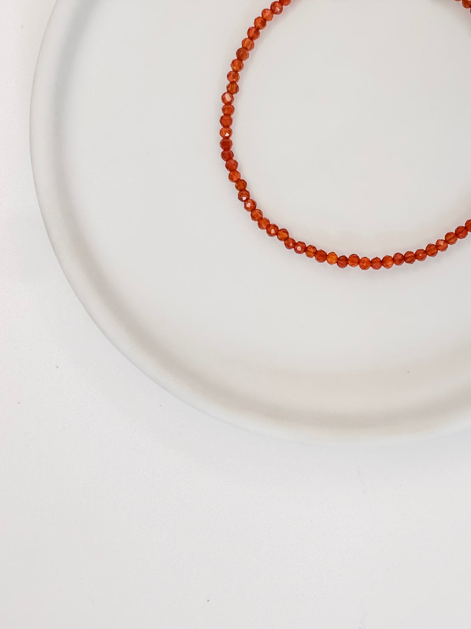 Pulseira em cristal natural vermelho Cornalina com esferas lapidadas de aproximadamente 2mm com fecho boia em prata, apoiada em bandeja de cerâmica branca e fundo branco.