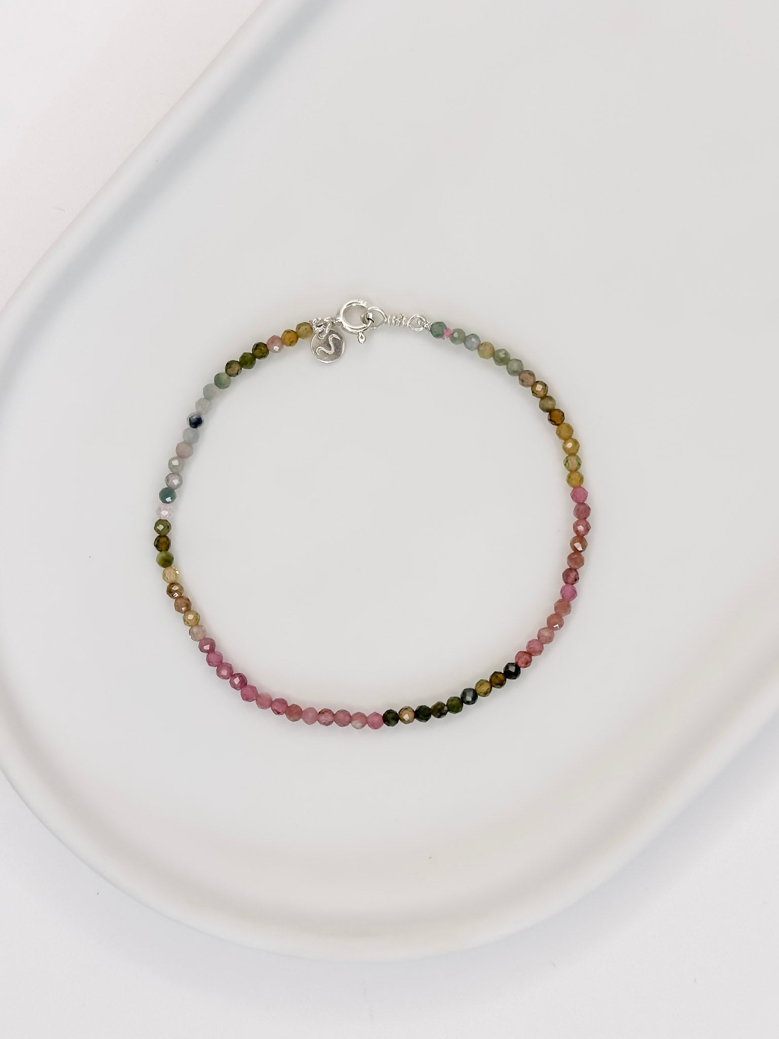 Pulseira ou tornozeleira com esferas lapidadas de aproximadamente 2mm de cristal natural turmalina melancia com fecho boia em prata apoiada em uma bandeja de cerâmica branca a fundo branco.