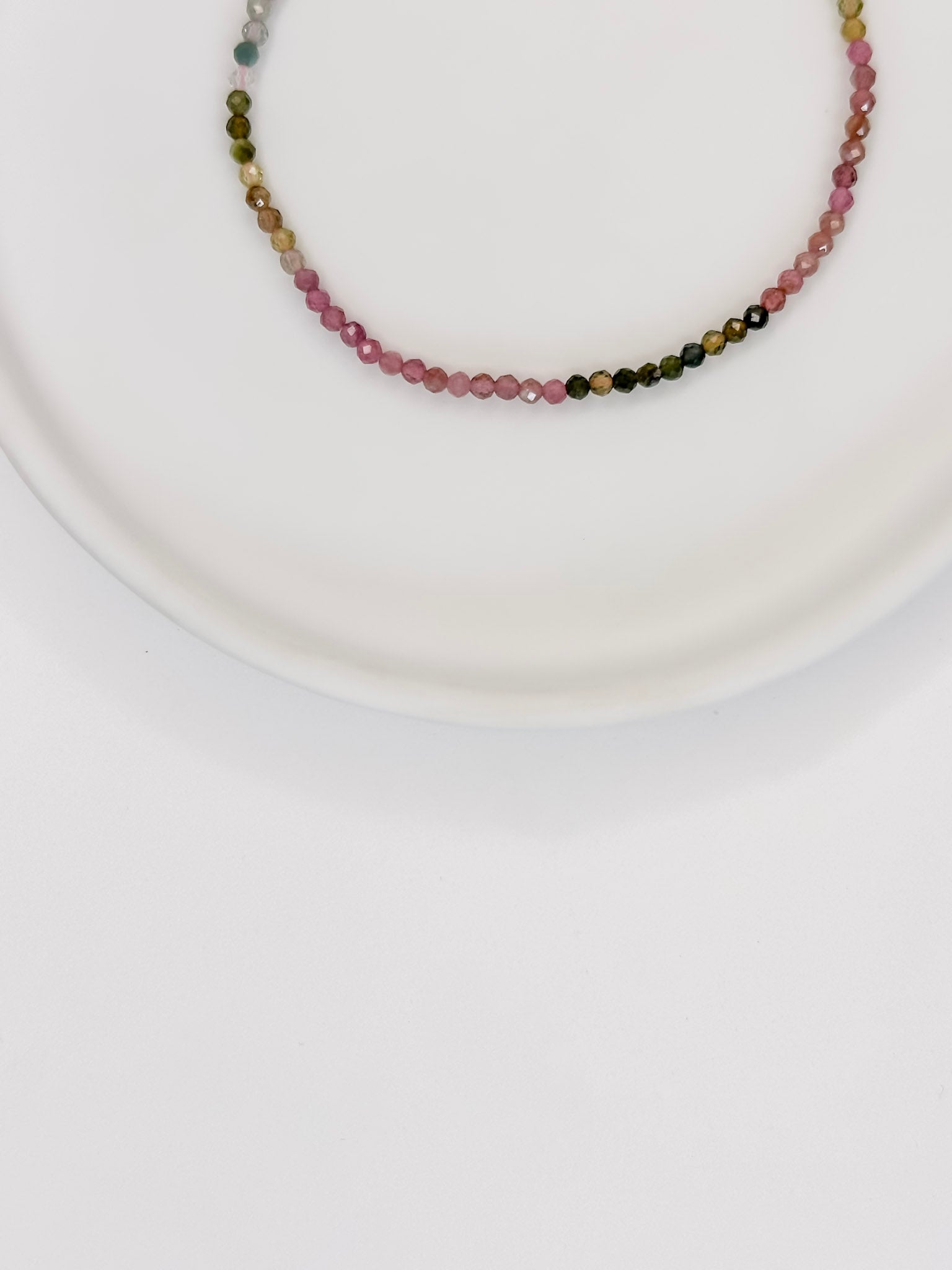 Pulseira com esferas lapidadas de aproximadamente 2mm de cristal natural turmalina melancia com fecho boia em prata apoiada em uma bandeja de cerâmica branca a fundo branco.
