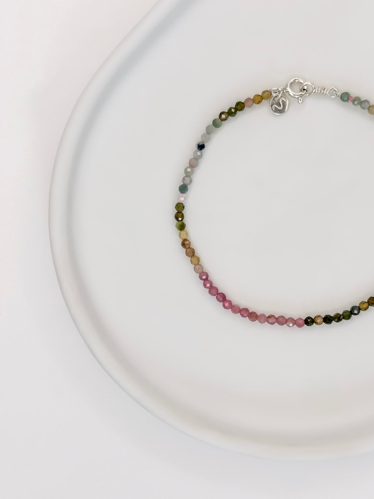 Pulseira com esferas lapidadas de aproximadamente 2mm de cristal natural turmalina melancia com fecho boia em prata apoiada em uma bandeja de cerâmica branca a fundo branco.