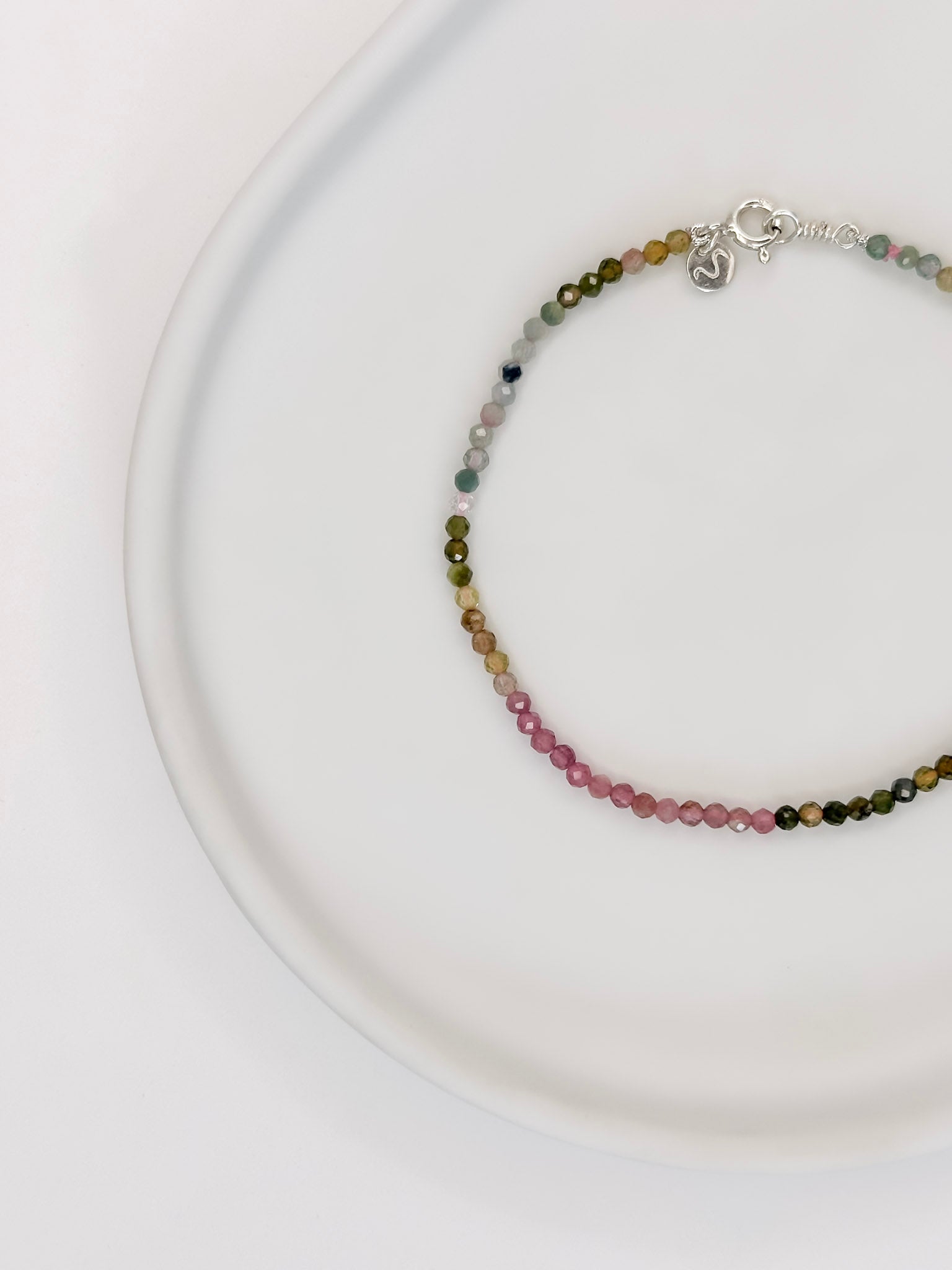 Pulseira com esferas lapidadas de aproximadamente 2mm de cristal natural turmalina melancia com fecho boia em prata apoiada em uma bandeja de cerâmica branca a fundo branco.