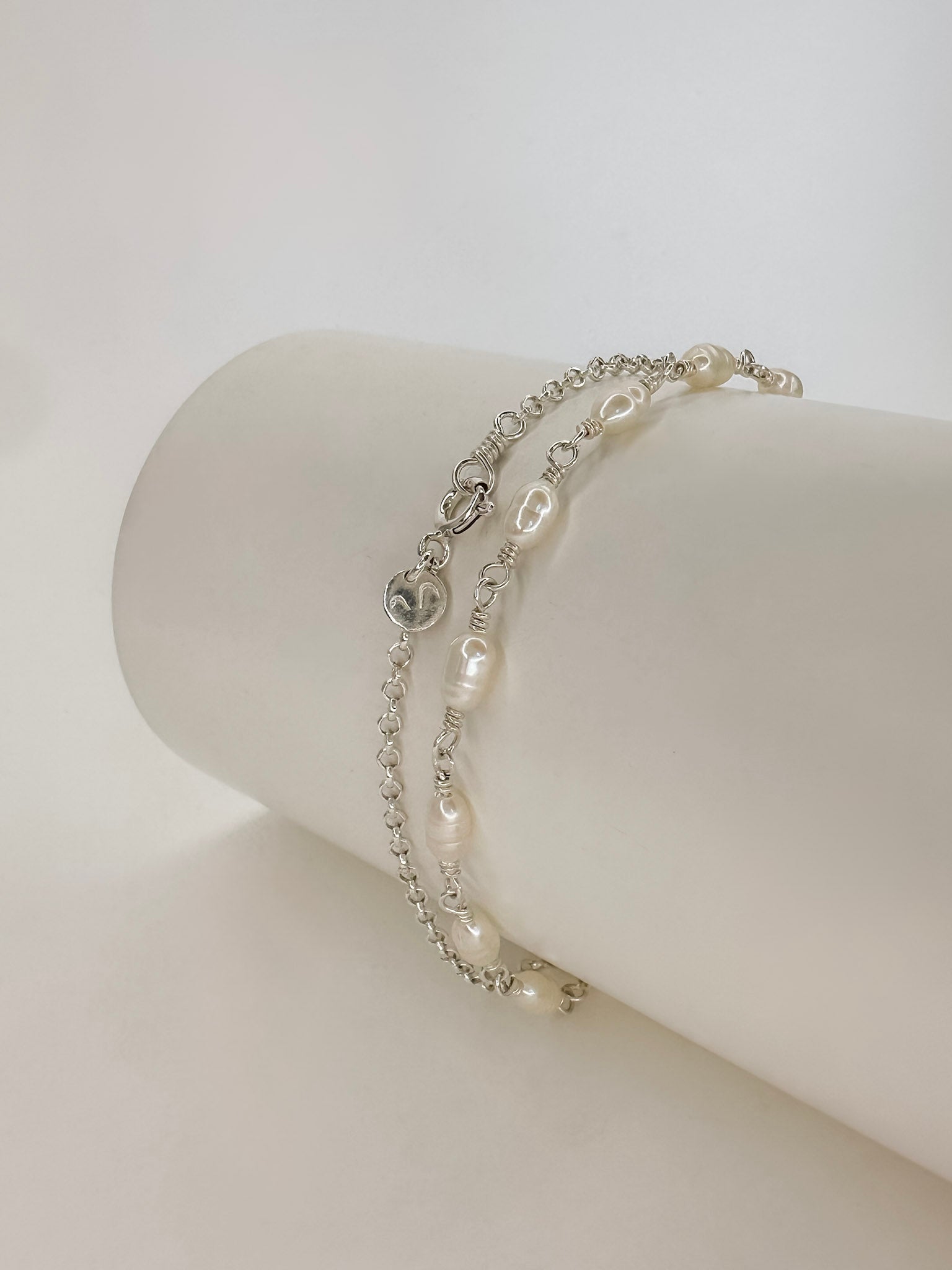 Pulseira e Colar de modelo choker feito em prata com pérolas naturais enrolado em um cilindro com fundo branco.