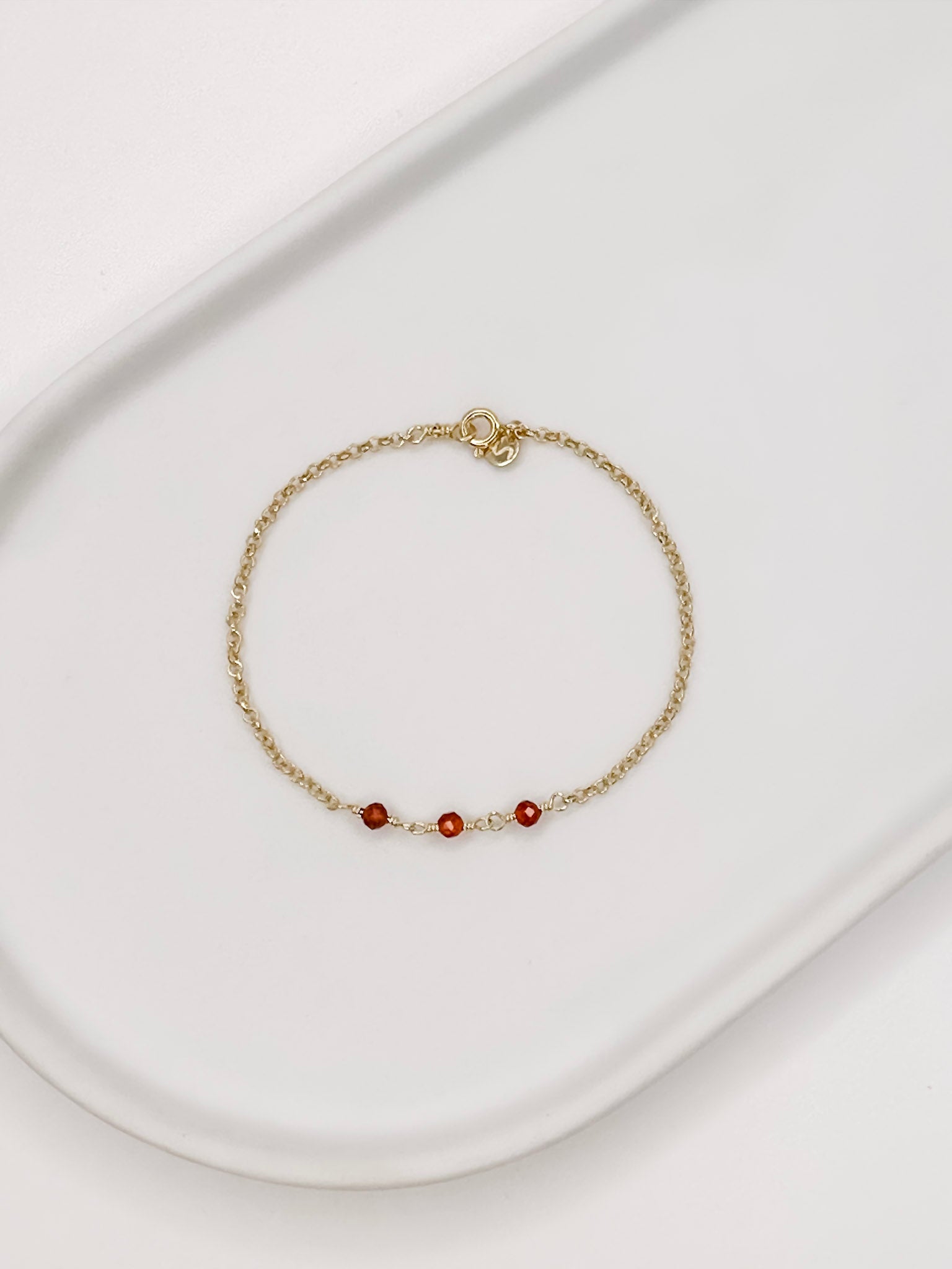 Pulseira em corrente portuguesa em prata com banho de ouro e 3 esferas de cristal natural Cornalina, apoiada em cerâmica branca com fundo branco.