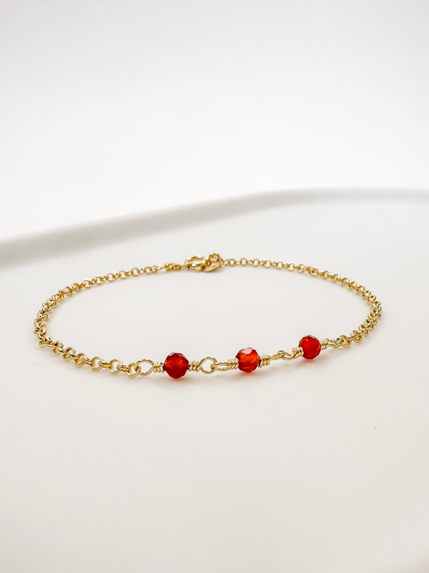Pulseira em corrente portuguesa em prata com banho de ouro e 3 esferas de cristal natural Cornalina, apoiada em cerâmica branca com fundo branco.