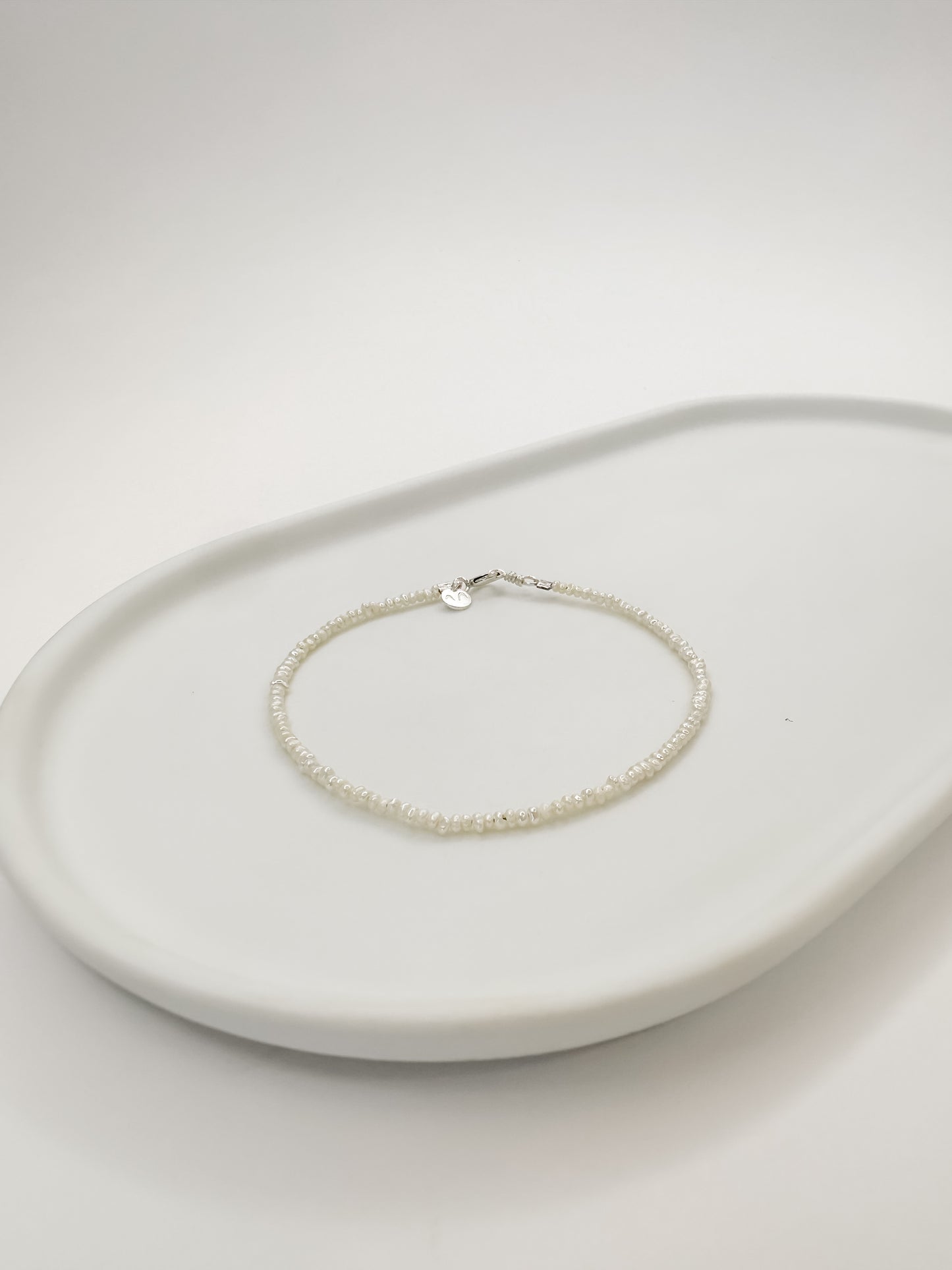 Pulseira de pérolas naturais de 2mm de medida cada, com fecho em prata, apoiada em uma bandeja de cerâmica branca com fundo branco. 